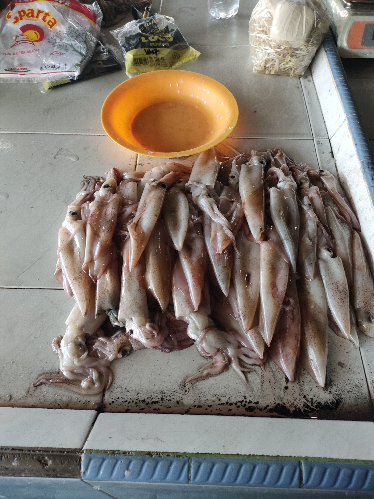 A close-up, overhead shot showcases a pile of fresh squid on a tiled surface, likely in a market stall. The squid are arranged in a natural, slightly overlapping fashion, with their bodies pointing upwards and tentacles fanned out. Their skin is a pale, translucent white with subtle pinkish-brown hues and speckled markings. Some squid appear to have their heads intact, with visible eyes. To the left of the squid, a bright orange plastic bowl filled with a light brown liquid is visible. Behind the bowl, two plastic bags are present: one white with red lettering reading "Sparta," and another yellow and black bag with Indonesian text. The tiled surface beneath the squid is grey and white, with some dark, smudged marks, suggesting it might be a workspace. The environment appears to be outdoors or in a covered market, given the lighting and the type of surface. The overall scene is one of a food market in Sungailiat, Indonesia, where fresh seafood is being prepared or displayed for sale.