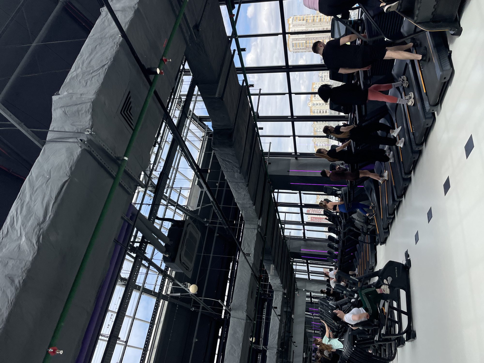 The image depicts a gym interior in Kazan, Russia. Rows of treadmills face a window wall showing a cityscape. People are exercising on the treadmills, mostly wearing athletic clothing. Overhead, exposed ductwork and support beams are visible against the sky. The floor is white with dark rectangular markings.