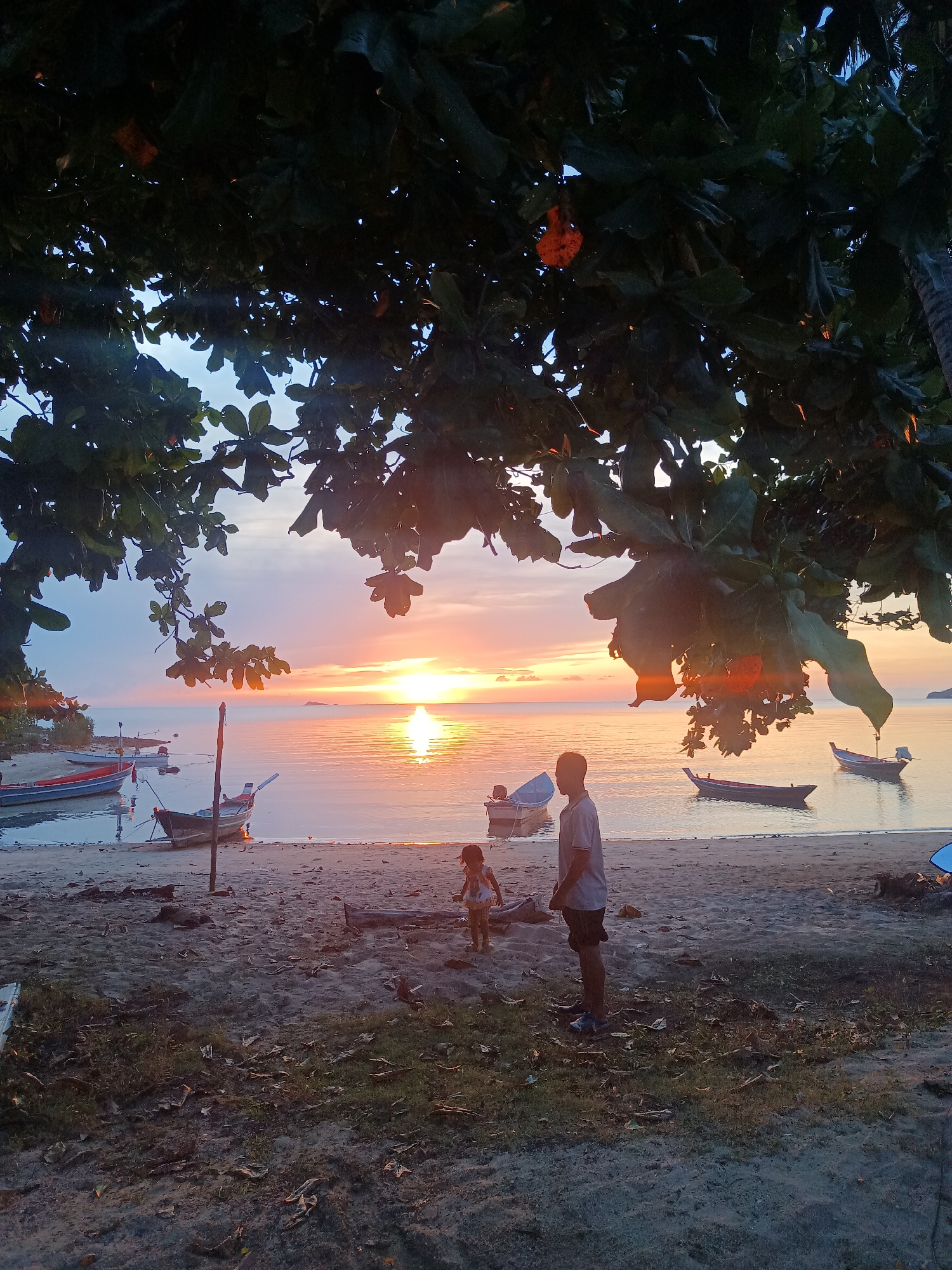 The image depicts a beach scene at sunset in Baan Wok Tum, Thailand. In the foreground, a sandy beach is partly covered with sparse vegetation and fallen leaves. A man stands on the beach, facing towards the sea and slightly to the right. Beside him, a child is also standing on the beach. The sunset in the background illuminates the water, creating a shimmering effect. Several small boats are moored in the shallow water along the shoreline. The scene is framed by the foliage of a large tree in the upper portion of the image.