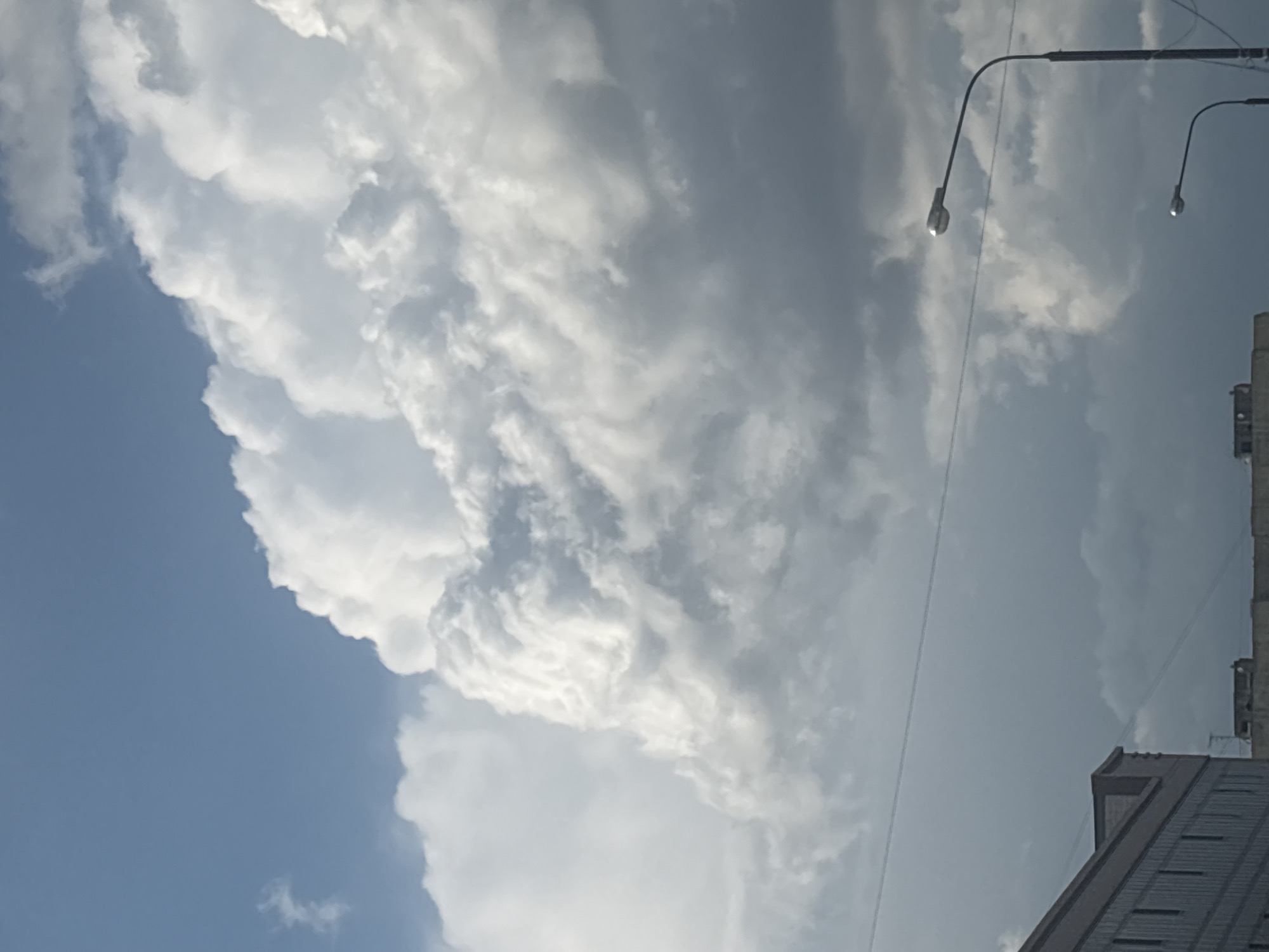 The image captures a dynamic sky over a city, likely Astrakhan, Russia, on a day with developing storm clouds. The upper portion of the frame is dominated by a vast, dramatic cumulus cloud formation, with bright white, towering masses interspersed with darker, more ominous grey areas indicating increased density and potential for precipitation. These clouds exhibit intricate patterns of swirling and texture, suggesting turbulent atmospheric conditions.
Below the dramatic cloudscape, a thin, horizontal line, possibly a power line or cable, stretches across the frame. Further down, the upper edges of urban structures are visible. On the left, a section of a building with a series of uniformly spaced windows is partially obscured. To the right, two streetlights with curved arms and distinct lamp heads are prominent. The lower streetlight is closer and more centered, while the upper one is positioned further to the right and higher. The background behind these structures is filled with the lower, darker segments of the cloudy sky. The overall impression is one of an approaching weather front over a metropolitan area.