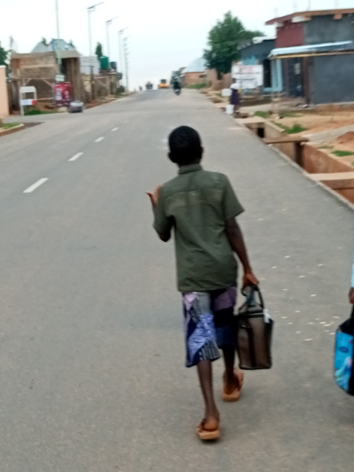 A young boy walks on a paved road, facing away from the camera. He wears a green short-sleeved shirt and patterned shorts, with light brown flip-flops on his feet. He carries a dark brown bag in his right hand and appears to be looking back, with his left hand raised. The road has white dashed lines marking the lanes and is bordered by a dirt path and some buildings. In the background, several vehicles and a person on a motorcycle are visible. The sky is overcast. The scene is set in Tempure, Nigeria.