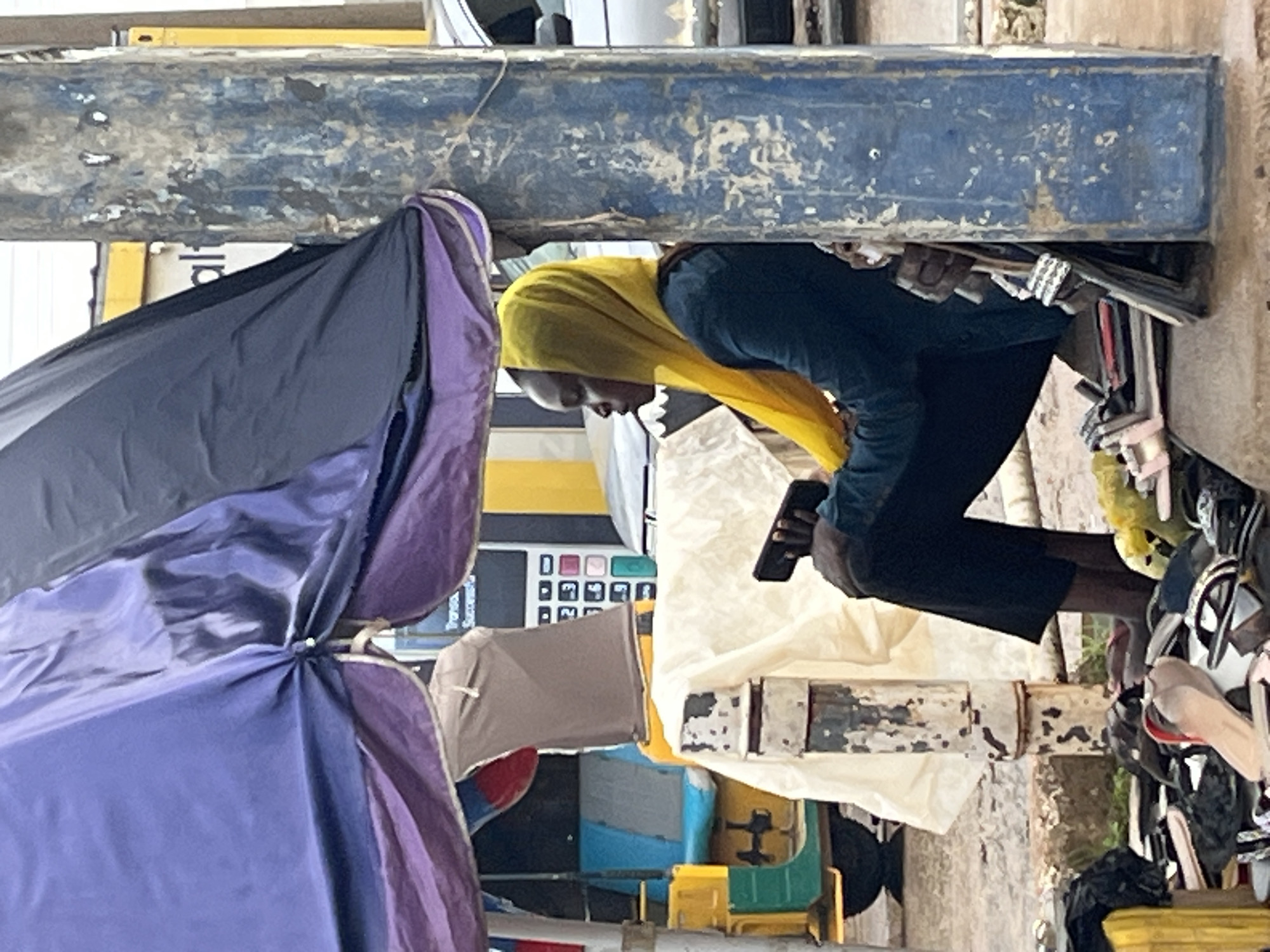 An overhead shot captures a street scene in Gura Top, Nigeria, focusing on a woman sitting under a canopy. The canopy, composed of purple and dark fabric, provides shade over a stall that displays various items. The woman, wearing a yellow headscarf and a dark blue jacket, is seated next to a thick, weathered blue pole. She holds a black mobile phone in her hands, her gaze directed forward. In front of her, a collection of footwear, primarily sandals, are laid out. To her left, partially visible, is a yellow and green motorized tricycle. In the background, a white calculator with large buttons and yellow signage is visible. The ground is paved, suggesting an urban setting.