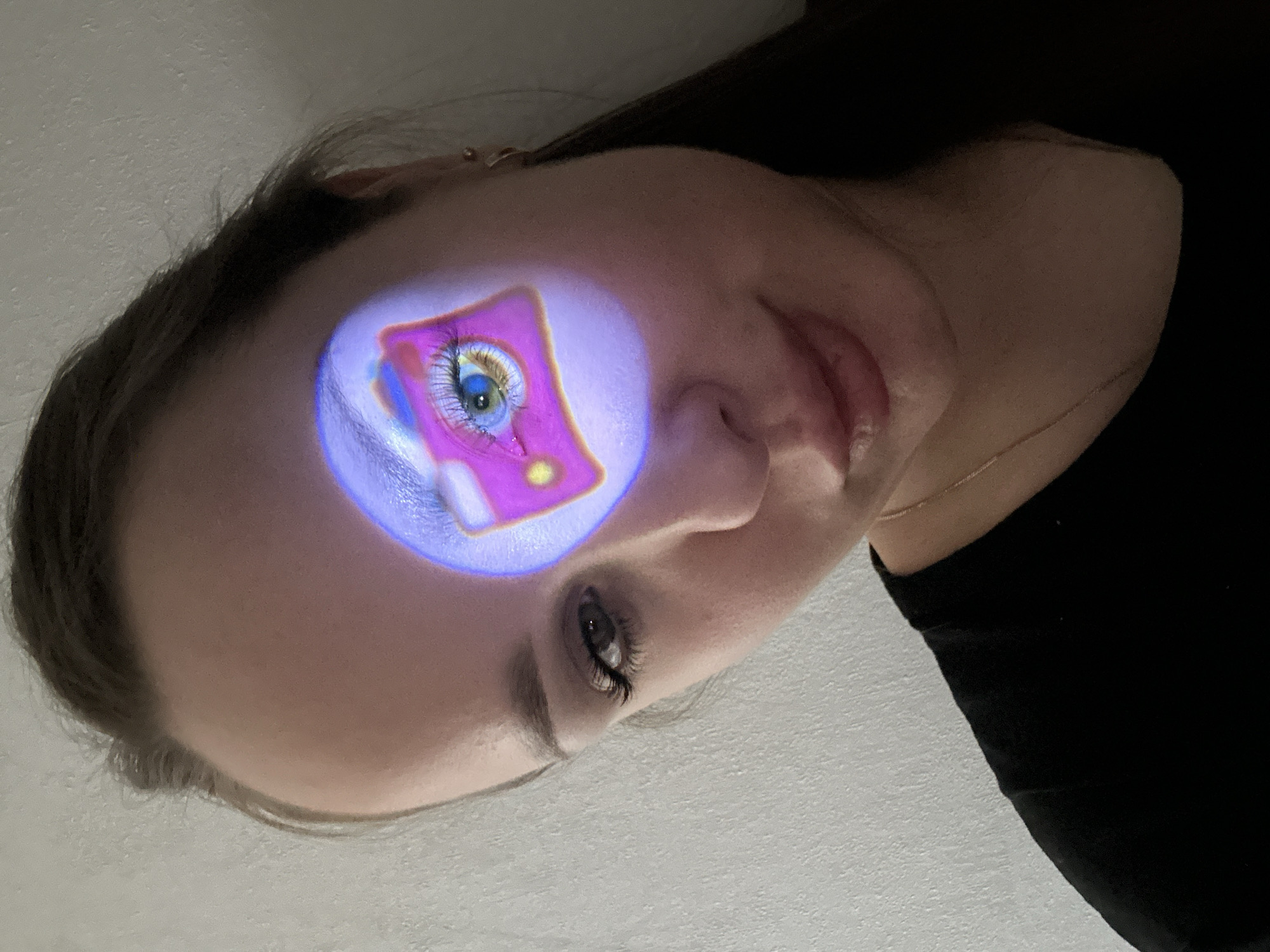 The media file displays a close-up, rotated portrait of a person, likely a woman, with an abstract, colorful image projected onto her forehead. The subject has fair skin, dark hair, and well-defined eyebrows. Her right eye (from the viewer's perspective) is partially visible, showing natural lashes and a dark iris, while her left eye is obscured by the projection. She wears a simple black top, and a delicate gold chain necklace is visible around her neck.
The central activity involves a bright, vibrant image being projected onto the person's forehead, creating a luminous, purple-hued glow on her skin. The projected image itself is a stylized representation of an eye, featuring long, exaggerated eyelashes, a striking blue iris, and a yellow pupil. This eye is framed within a prominent, neon pink or magenta square shape, which includes some smaller circular yellow details. The colors of the projection are vivid and contrasted against the person's natural skin tone.
Notable details include the crisp focus on the subject's face and the projected image. The person's expression is neutral and calm, suggesting a posed photograph. The background is a plain, light-colored, slightly textured surface, possibly a wall or ceiling, indicating an indoor setting. The time of day is not discernible, as the scene appears to be lit artificially to facilitate the projection. No text is visible in the image. The provided context indicates the location is Chaikovsky, Russia, though this is not visually evident in the media.