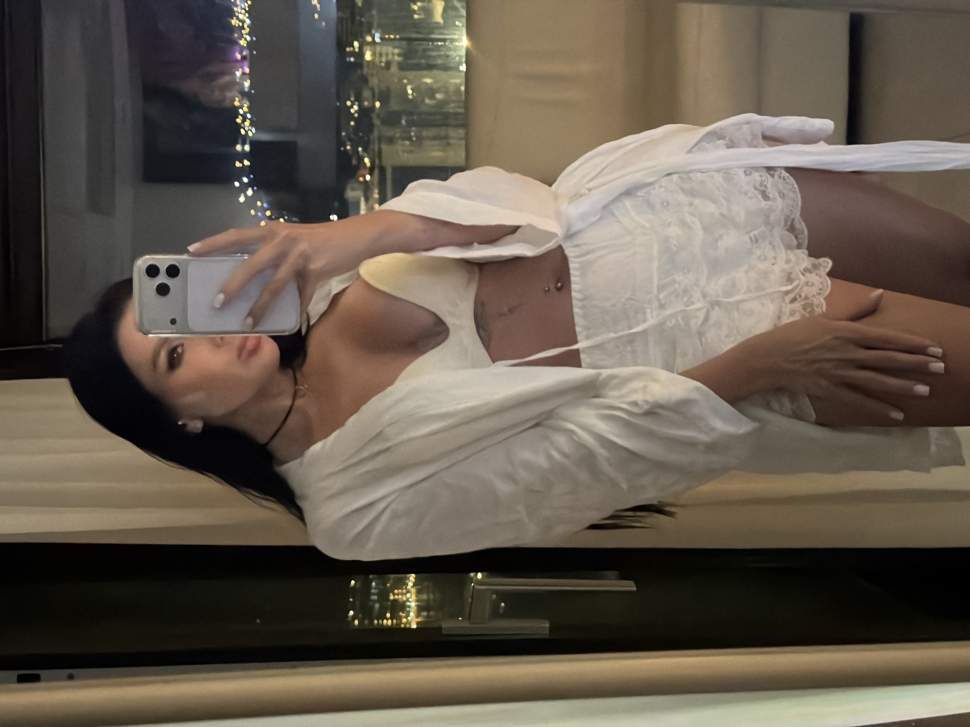 A forensic examination of the image reveals a lone female subject situated indoors, specifically within a room overlooking the city of Boreti, Montenegro, at night. The subject, possessing long dark hair, is captured from the upper torso to the mid-thigh, engaging in a mirror selfie. She is clad in a loose, white, open-front robe, a light-colored brassiere, and white lace-trimmed shorts. Accessories include a dark choker necklace with a small pendant and a double-ball belly button piercing. Her fingernails exhibit a white polish. She holds a light-colored smartphone, equipped with a three-lens camera system, which obscures the central portion of her face. Her left hand rests on her upper thigh. The background features a large window revealing a blurred nocturnal cityscape with numerous illuminated structures. To the immediate left, a light-colored curtain and a dark-framed glass door with a metallic handle are discernible. The indoor environment is dimly lit, contrasting with the vibrant outdoor city lights.