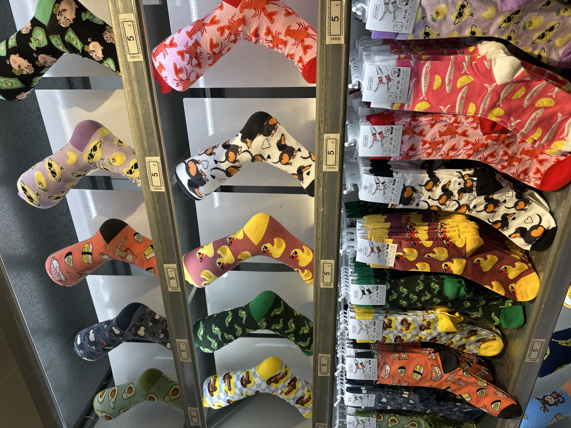 The image shows a retail display of novelty socks. The socks are presented on a multi-tiered shelf system, with some displayed on mannequins and others hanging from hooks. The socks feature various colorful patterns, including avocados, sushi, dogs, ducks, lobsters, fish, and pigs. Several price tags, indicating "5 €," are visible on the shelves. The overall environment appears to be an indoor store. There are no people visible in the image.