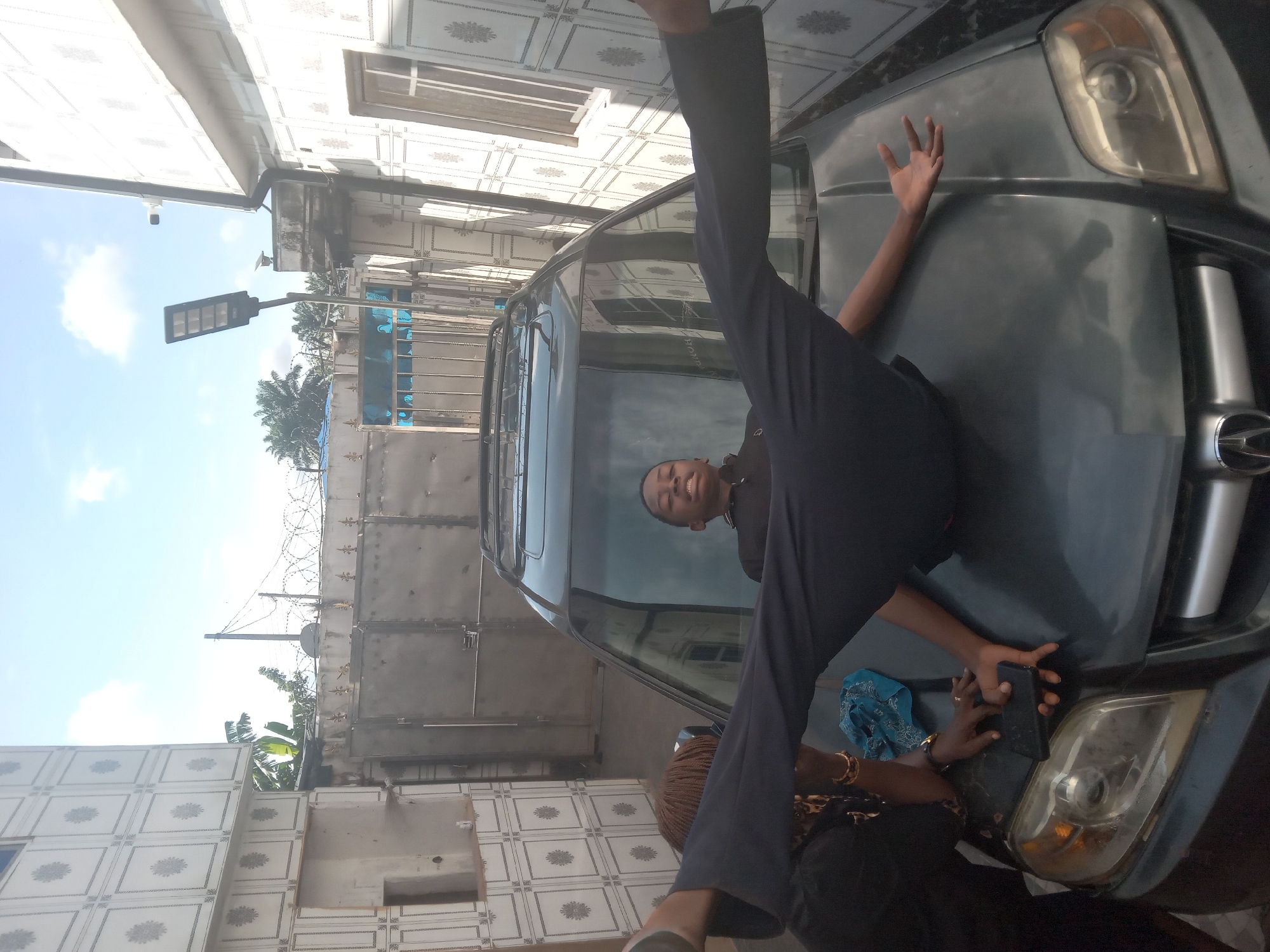 The image, correctly oriented, displays an outdoor scene in Owa-Alero, Nigeria, during daytime under a clear, bright sky with some visible clouds.
The central subject is a dark-colored SUV, likely a Toyota, occupying the foreground. A young person, possibly male, is dramatically posed on the car's hood. His body is stretched out, with one leg extended high into the air and the other bent, creating a wide V-shape. His head and torso are reflected in the car's windshield, revealing a smiling face looking towards the viewer. He is wearing dark trousers and a dark top.
Near the front bumper and headlight of the car, another person's hands are visible, holding a black smartphone. This person, whose face is not visible, appears to be operating the phone, possibly capturing the moment of the pose on the car. A blue piece of fabric is also seen near their hands.
The background features a building on the left with a distinctive white tiled exterior, adorned with a repeated patterned motif. On the right, a tall metallic gate or fence, topped with barbed wire, separates the foreground from further background elements. Behind the fence, several palm trees and other tropical foliage are visible. A modern-looking LED streetlight on a pole stands prominently in the middle-ground, indicating contemporary infrastructure.
The overall mood appears lighthearted and playful, with the person on the car smiling. The bright sunlight suggests good weather conditions. No explicit text is clearly legible, though the car's emblem is faintly visible.