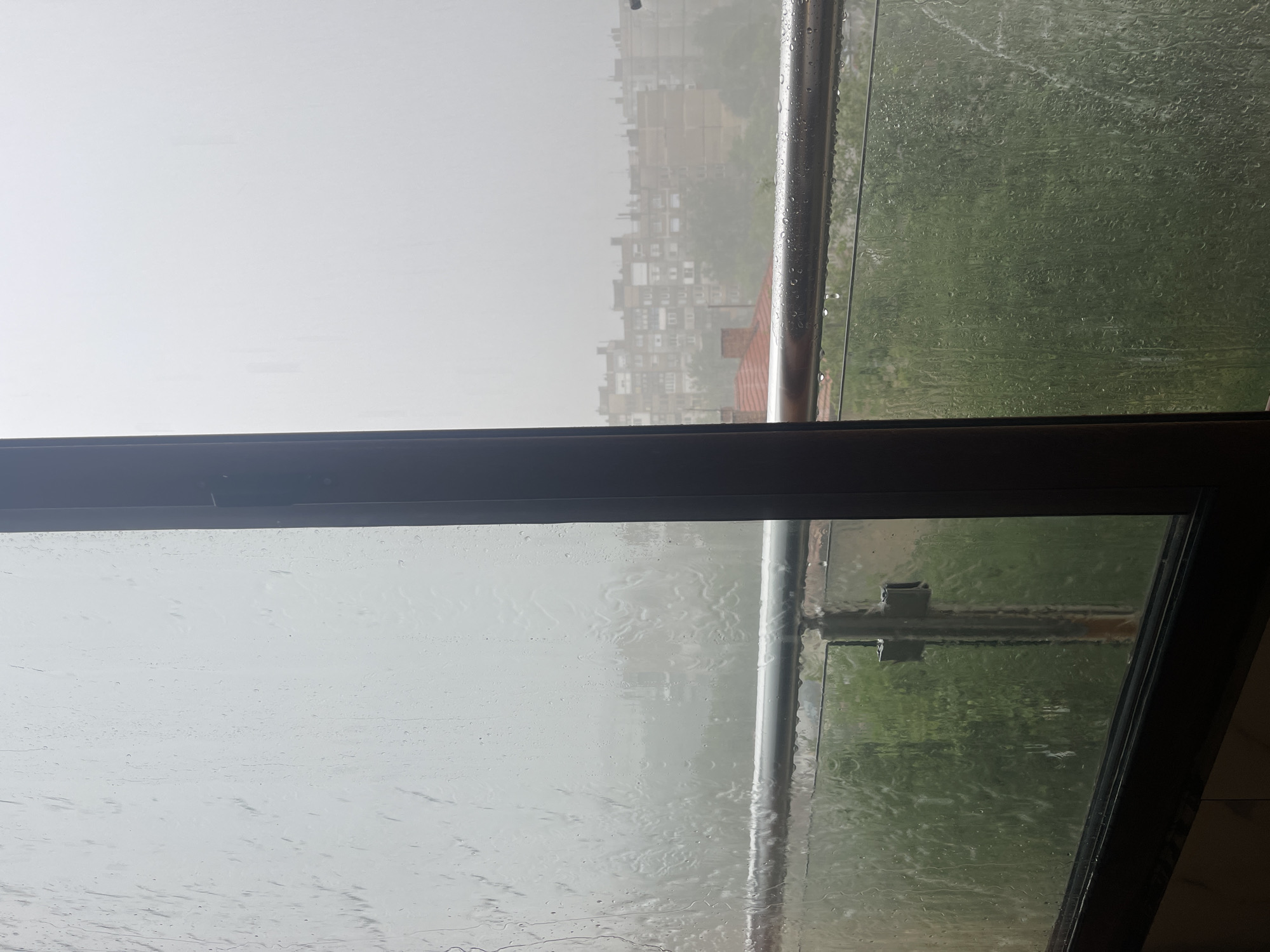 The image captures a view through a rain-streaked window or glass enclosure on an overcast day. The perspective is from indoors or a covered balcony, looking outwards.
In the foreground, the glass surface is heavily covered with numerous water droplets and streaks of rain, distorting the view beyond. A dark, possibly wooden or metal, frame runs along the bottom, resembling a window sill or part of a balcony railing structure. To the right, a vertical dark frame or divider is visible, along with a metallic cylindrical railing that is also speckled with water droplets.
Through the watery glass, the midground reveals a blurred urban landscape. Buildings, likely residential, with multiple windows and balconies, are visible but appear indistinct due to the rain and atmospheric haze. The colors are muted, and the overall visibility is low. To the far right, beyond the metal railing and through more rain-covered glass, there are hints of green foliage, suggesting trees or grass.
The scene depicts heavy rainfall and an overcast sky, with the light appearing diffused and soft. No people or specific activities are visible. There is no discernible text in the image. The general impression is one of a quiet, rainy day viewed from a sheltered position.
