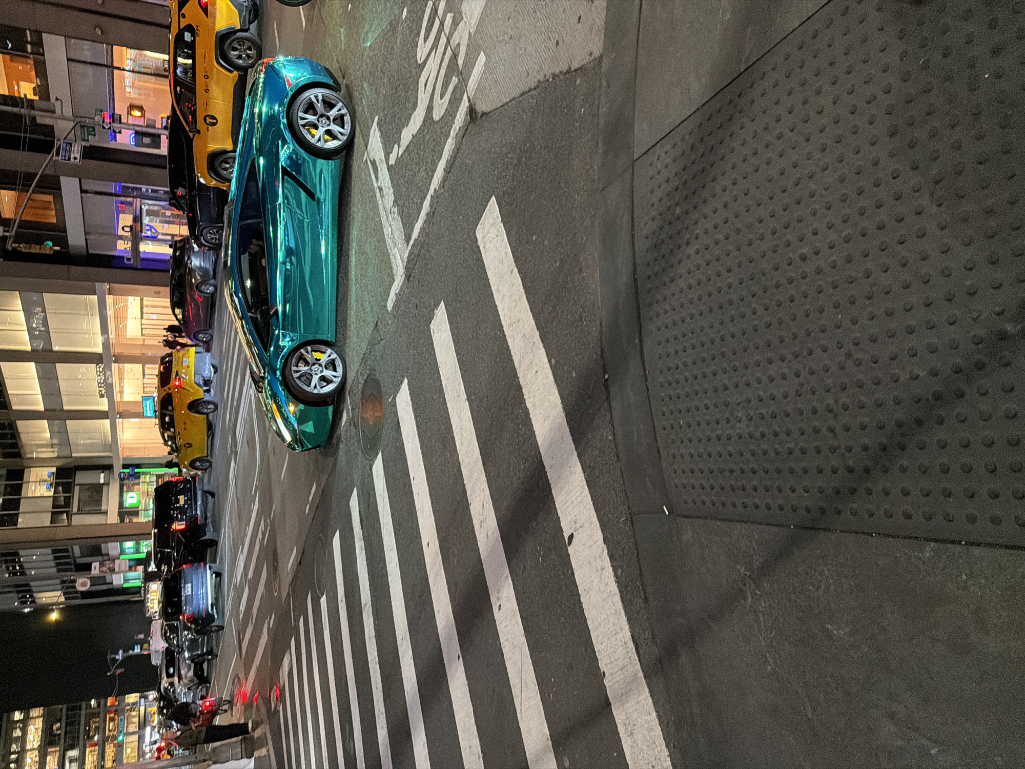 This image, captured at night, depicts a vibrant urban street scene, oriented 90 degrees clockwise. A striking metallic teal or turquoise sports car, likely a Lamborghini, is prominently featured in the foreground, facing right, with its shiny surface reflecting the city lights. Its silver multi-spoke wheels reveal bright yellow brake calipers.
Behind the sports car, a line of vehicles is visible. This includes at least two distinctive yellow New York City taxis; one bears the number "T734". Other assorted passenger cars, including sedans and SUVs, are also in the queue or parked along the street. Further down the street, blurred by distance and motion, more vehicles and several pedestrians can be discerned on the sidewalk.
The street itself features clear white crosswalk lines stretching across the asphalt. Near the top of the image, painted on the street, partially visible white text spells out "NO STOPPING" or similar traffic instructions. To the right of the street, a dark grey sidewalk section includes a tactile paving strip with raised dots, indicating accessibility features.
In the background, modern buildings with large, reflective glass windows rise, illuminated by interior lights and street lamps, casting reflections onto the cars and street. Traffic lights and other street signage are also visible. The overall scene suggests a busy night in a major city, confirmed by the presence of yellow taxis as New York City. The weather appears clear.