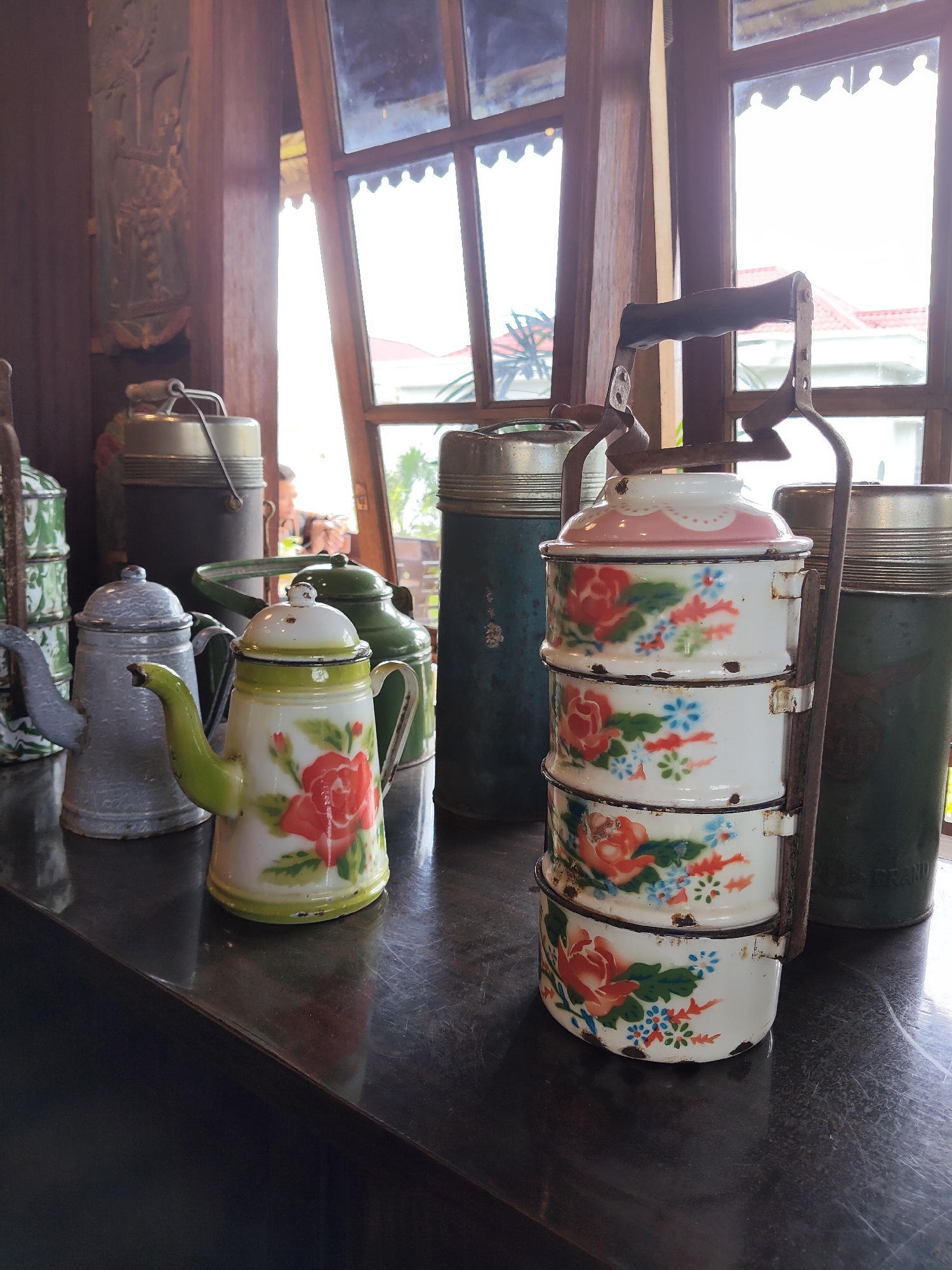 A close-up, eye-level shot features a collection of vintage enamelware objects displayed on a dark, reflective surface. In the foreground, a white and green enamel teapot adorned with red roses and blue flowers is prominently positioned. To its left is a gray enamel teapot with a dark spout. Behind the teapots, a tall, dark green enamel cylinder with a lid stands. To the right of the teapots, a white, three-tiered enamel food carrier, also decorated with red roses and blue flowers, is held together by a metal frame. Further to the right is another tall, dark green enamel cylinder. The background reveals a window with multiple panes, through which sunlight streams in, illuminating a hint of red-tiled roofs and foliage outside. The overall aesthetic suggests a display of antique kitchenware, possibly in a shop or home in Medan, Indonesia.