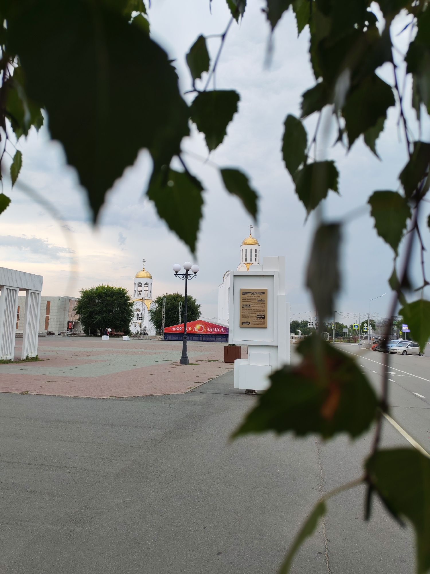 In an outdoor urban setting, a white and gold domed church with a cross on top is visible in the background. In front of the church, there is a paved area with red and grey paving stones, a street lamp with three lights, and some trees. To the right of the paved area is a white, modern-looking structure with a plaque on it. A street with cars is also visible to the right of the scene. The foreground is partially obscured by out-of-focus green leaves. The sky is overcast.