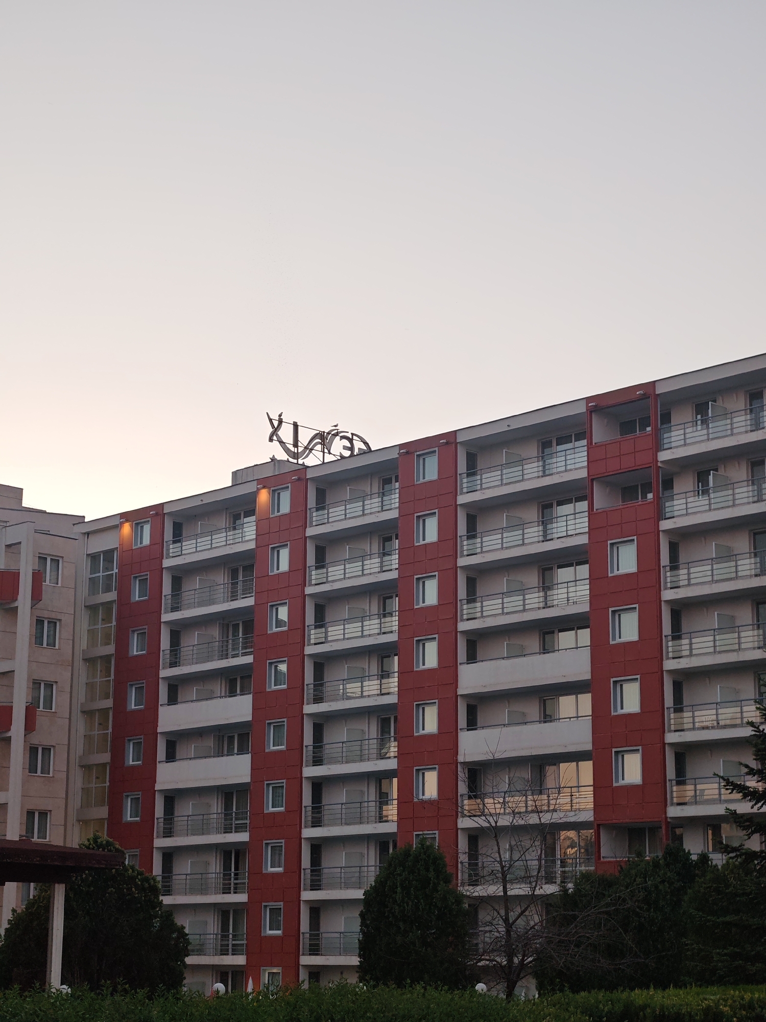 A daytime exterior shot of a tall, multi-story building in Nesebar, Bulgaria. The building features a red and white facade with balconies and rectangular windows. The top of the building is adorned with a metallic, abstract, cursive design, possibly a name or logo. The sky is a pale gradient of color. Green bushes and trees are visible at the base of the building.