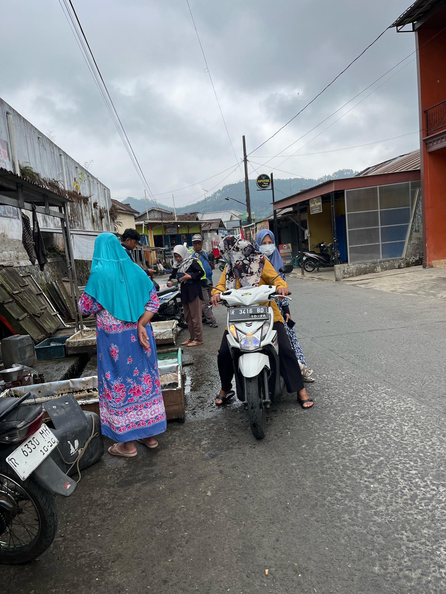 In Karangkobar, Indonesia, on a cloudy day, two women are riding a white scooter on a wet asphalt road. The woman in front wears a yellow long-sleeved shirt and black pants, with a floral-patterned headscarf covering her hair and a surgical mask over her mouth and nose. The woman behind her wears a blue headscarf and a blue and pink floral patterned dress. She is also wearing sandals.
To the left of the scooter, a woman in a blue headscarf and a blue dress with pink floral patterns stands facing away from the camera, her back to the scooter. She is wearing light brown sandals. In the background, there are several buildings, including a structure with a red-orange facade and a darker tiled roof. Other buildings appear to be shops or market stalls with corrugated metal roofs. A sign on a pole reads "APOTEK BUANA" with a logo. The road is lined with wooden stalls or market stands, some containing what appear to be crates and other items. The ground is wet, suggesting recent rain. A motorcycle license plate is visible on a black box attached to the scooter, reading "R 3416 BD". The overall scene depicts a street market or a bustling commercial area.