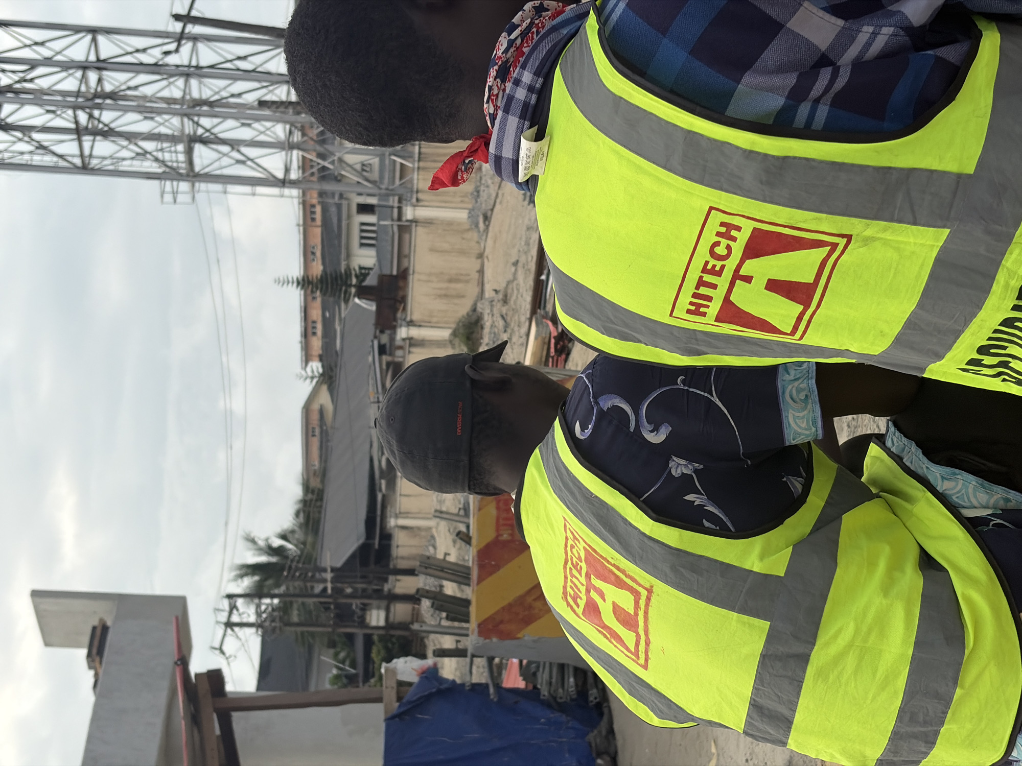 Two individuals are observed from behind, positioned outdoors in what appears to be a construction or industrial area in Alaguntan, Nigeria. Both individuals are wearing high-visibility neon yellow vests with reflective gray stripes, indicating they are likely workers or security personnel. The person on the left is wearing a dark baseball cap and a dark patterned shirt underneath the vest. The person on the right is wearing a red, white, and blue bandana around their neck and a blue and black plaid shirt. Both vests feature a red rectangular emblem with the word "HITECH" above a stylized "A" symbol. In the background, there is a partially constructed concrete structure, scaffolding, metal towers, buildings with visible rooftops, and overcast sky. The ground is uneven and appears to be dirt or gravel.