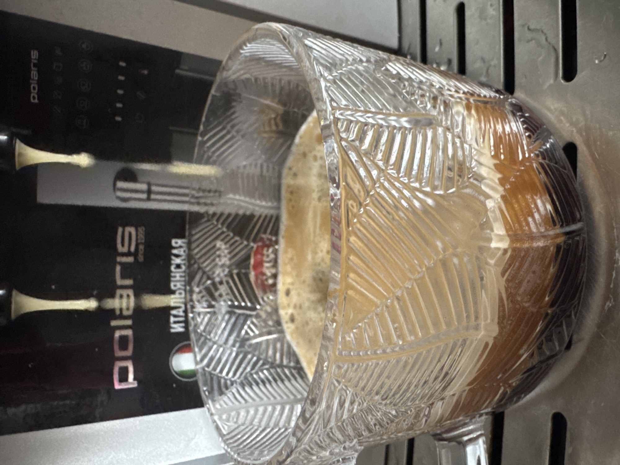 A glass mug with a decorative, faceted surface contains a beverage with a frothy top and a dark liquid below. The mug is placed on a metal surface next to a Polaris-branded coffee maker, which includes the words "ITALIYANSA" and an Italian flag. It is not possible to definitively confirm if this image was taken in the city of Osa, Russia, based on the provided information.