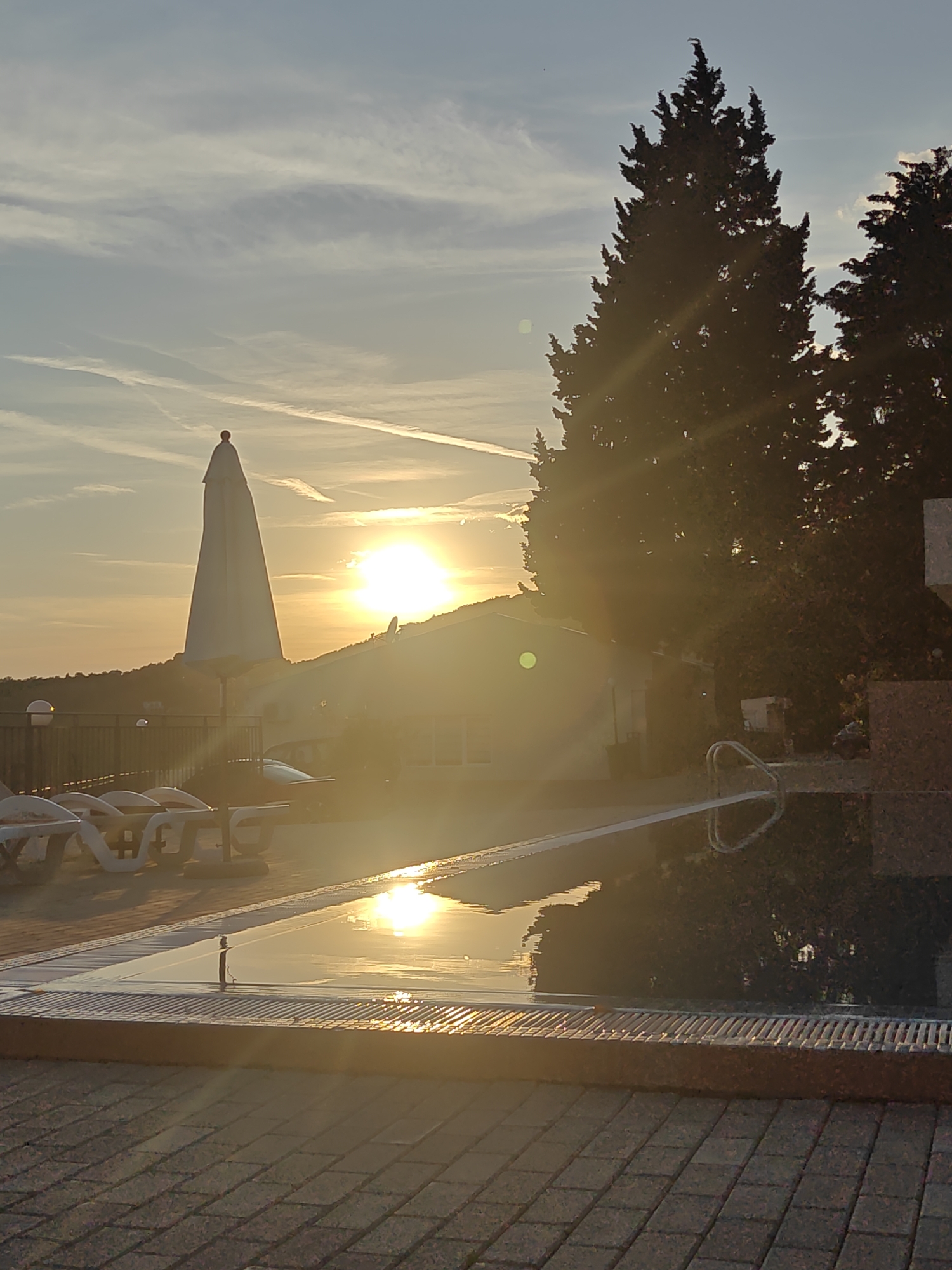 The image depicts a tranquil sunset scene in Šušanj, Montenegro, likely taken at a resort or private residence. In the foreground, a tiled patio leads to a swimming pool, its surface reflecting the intense sunlight and sky. Several lounge chairs are lined up near the pool, hinting at relaxation and leisure. In the background, a structure is partially obscured by the bright sunlight, positioned on a slight elevation, with evergreen trees flanking it. A white parasol adds a geometric element to the scene. The sky is a mix of light blue and yellow hues with wispy clouds and contrails, indicating the passage of aircraft. The overall atmosphere is warm and peaceful, characteristic of a coastal sunset.
