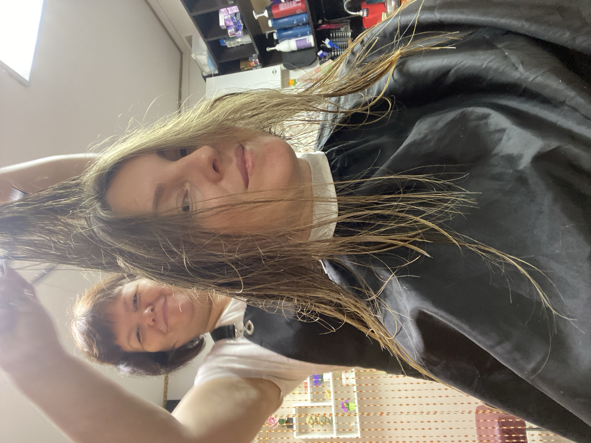 A middle-aged woman with wet, blonde hair, wearing a black salon cape, is having her hair combed by a shorter woman with brown hair and a white t-shirt in a salon. Shelves containing hair products and other beauty supplies are visible in the background. The location is Noviy, Russia.