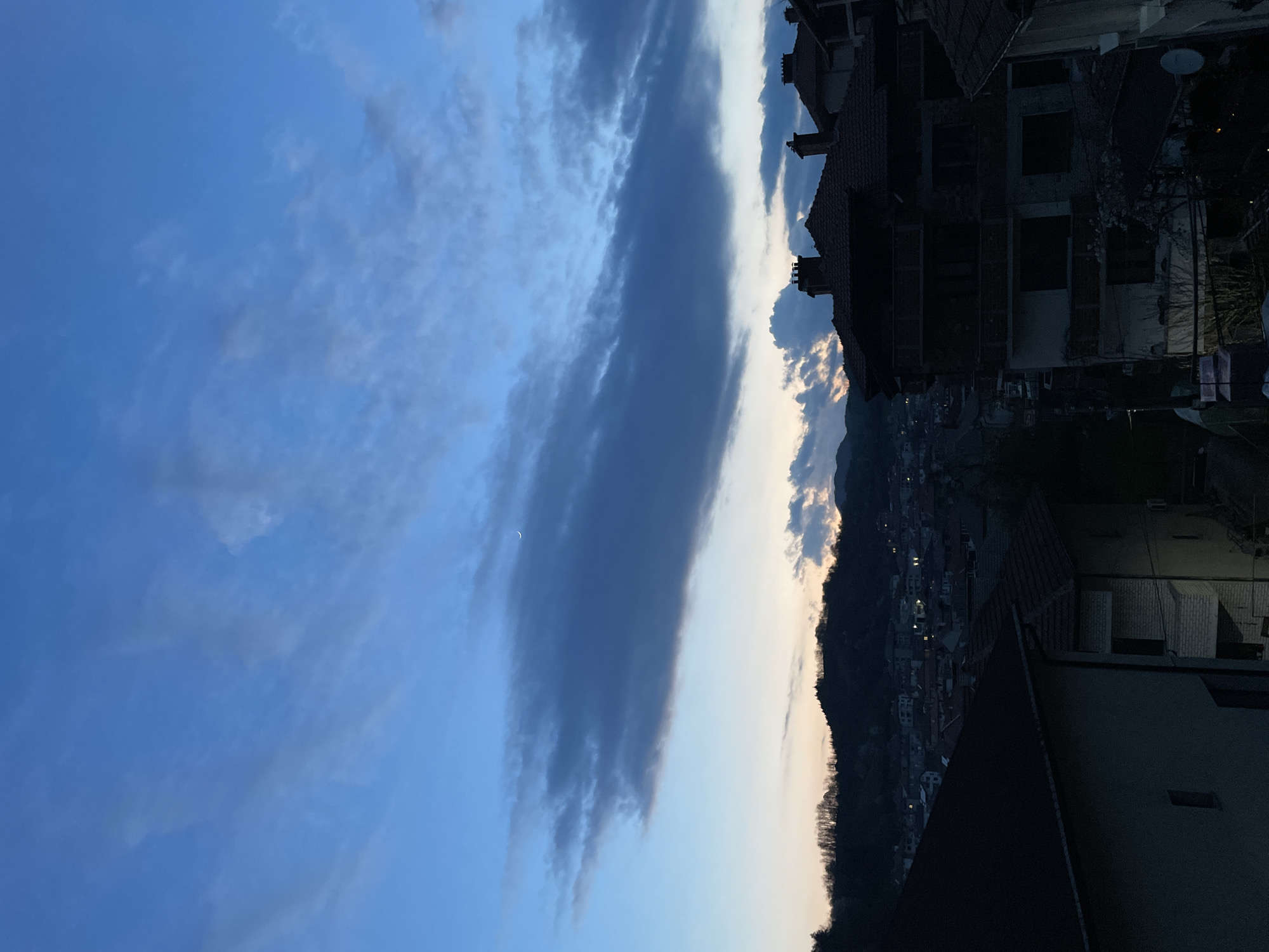 The image depicts a twilight scene in Zlatograd, Bulgaria. The sky is predominantly blue with a band of dark, elongated clouds stretching vertically across the frame, and a sliver of crescent moon visible. Silhouetted buildings with red tiled roofs are on the right. In the middle ground, a dark, tree-covered hillside gives way to a cluster of lit buildings, likely the city center. The foreground shows rooftops and other parts of buildings with limited detail. The image is taken from an elevated position looking towards the west/southwest.