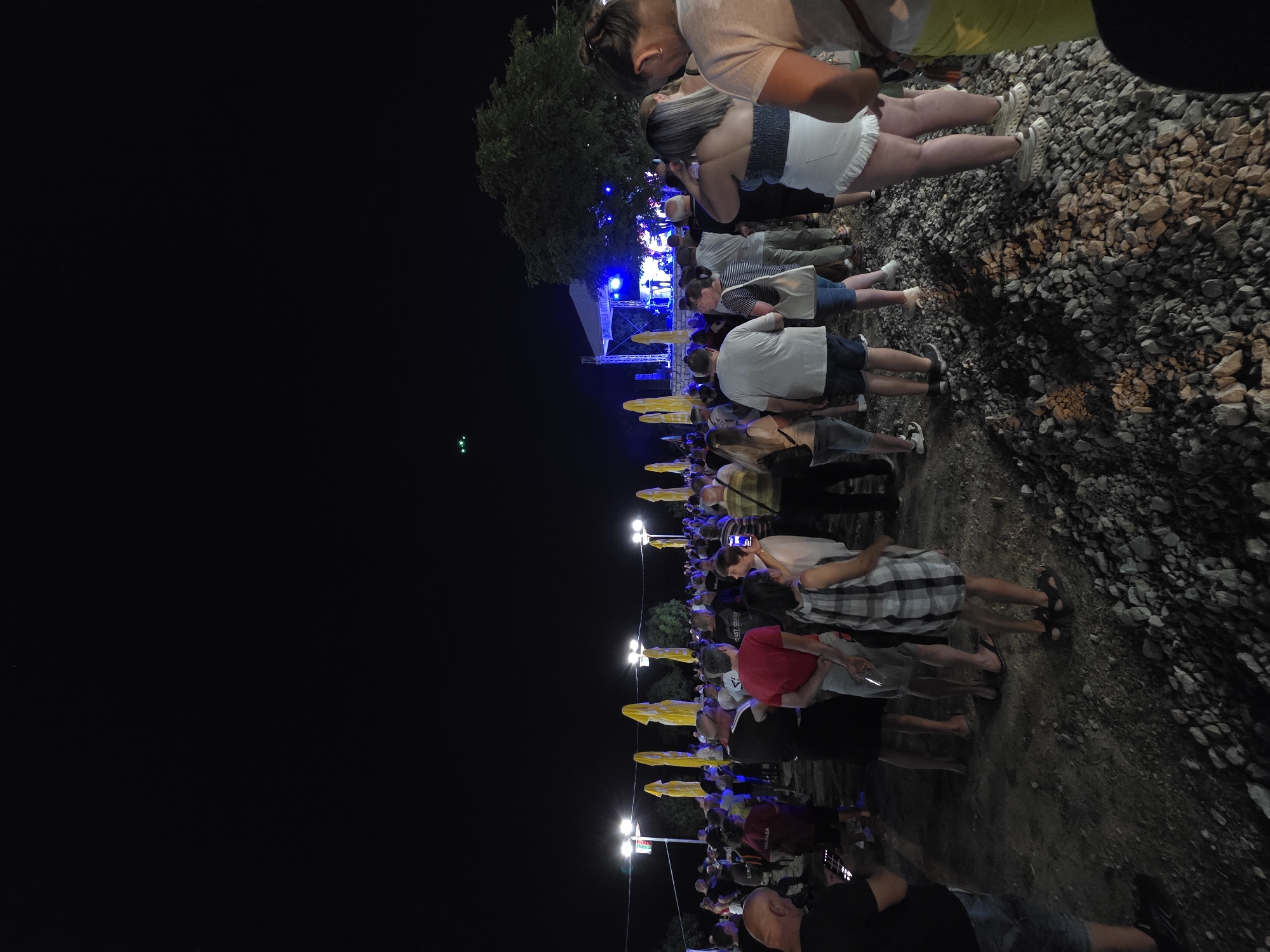 This is a night scene captured from a low angle, looking towards a stage at an outdoor event. The foreground is dominated by a pile of rough stones and gravel. In the midground, a crowd of people are gathered, facing towards the stage. They are dressed in casual summer attire. Behind the crowd, several tall, bright yellow umbrella-like structures are visible, illuminated by lights. Further back, a stage is set up with a band performing under blue lights. The background consists of a dark sky, with a few trees visible to the right of the stage. The overall atmosphere suggests a lively outdoor concert or festival. The location is identified as Gara Oreshets, Bulgaria.