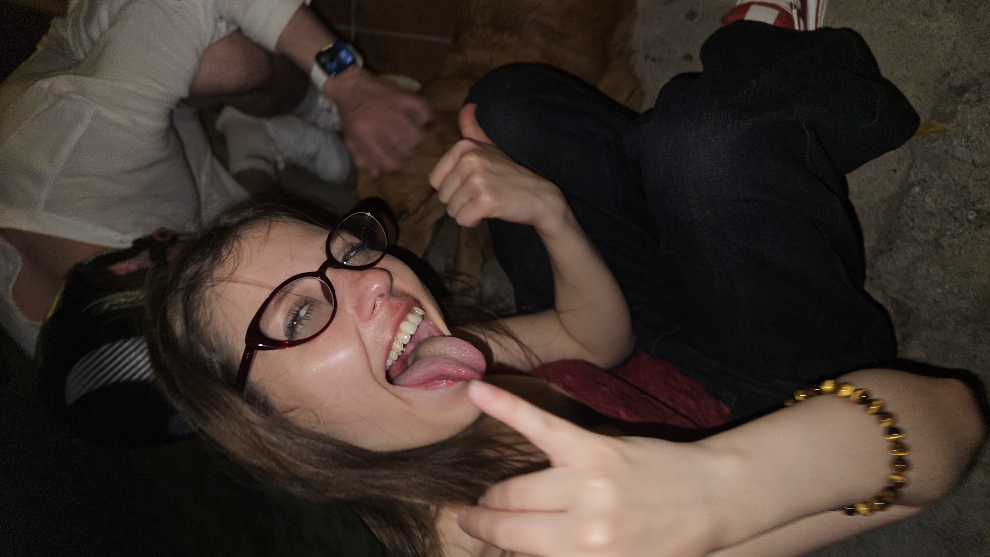 A woman wearing glasses is lying down with her tongue out, making a hand gesture. She is wearing a reddish top and black pants. A bracelet is on her wrist. Another person is visible behind her, wearing a white shirt and a smartwatch. The setting appears to be indoors, possibly on a concrete floor. There's no information about Nha Trang, Vietnam within this image.