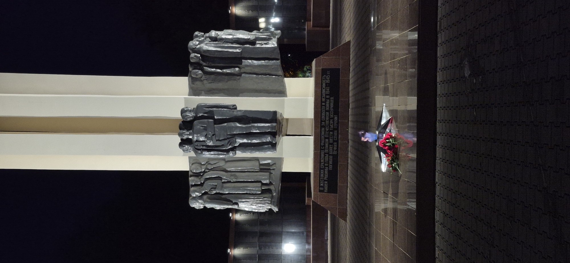 This media file depicts a monument at night, likely a memorial dedicated to soldiers. The monument features several tiers of sculptures, each depicting a group of figures lying down, possibly representing fallen soldiers. The sculptures are dark and appear to be made of metal or stone.
The scene is illuminated by artificial lighting, casting shadows and highlighting the textures of the monument and its surroundings. To the right of the monument, a wall is visible, adorned with a star-shaped emblem and a bouquet of red flowers, suggesting a memorial for a military event.
There is visible text inscribed on a dark plaque on the wall. The text is in Russian and appears to be a dedication, mentioning "cruel battles with fascism," "our Motherland," "the Great Patriotic War," and "more than six thousand Ussuriysk residents" who perished. It also includes the phrase "Eternal glory to the heroes!"
The overall atmosphere is somber and commemorative, appropriate for a memorial site. The time of day is clearly nighttime, and the setting appears to be an outdoor public space. The mention of "Ussuriysk" in the text indicates the location is Ussurijsk, Russia. The weather cannot be determined from the image.