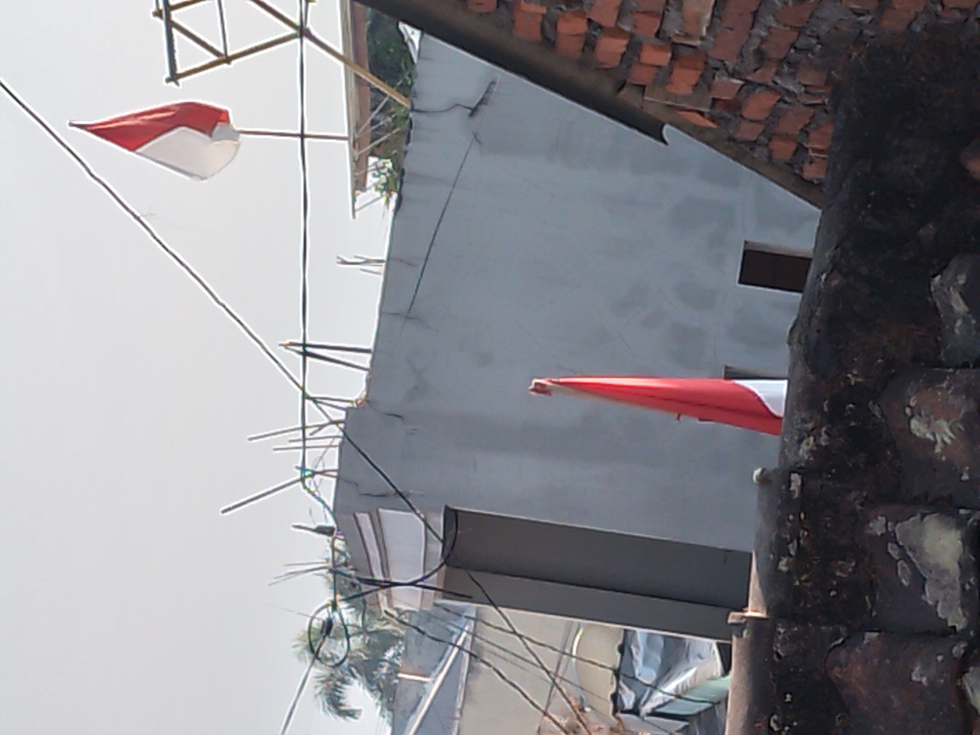 In the city of Batu, Indonesia, two Indonesian flags, red and white, are displayed outdoors. One flag is smaller and attached to a pole, positioned above and to the right of a building with a grey, textured wall. This flag is partially obscured by wires and appears to be flapping in the wind, showing its two distinct colors. The second flag is larger and prominently displayed on a pole near the center of the image, positioned on what appears to be a rooftop. It is also red and white and hangs vertically. The surrounding environment consists of rooftops, a portion of a brick wall, trees, and numerous electrical wires. The sky is a pale, overcast white. The scene suggests an outdoor setting with a focus on national symbols.