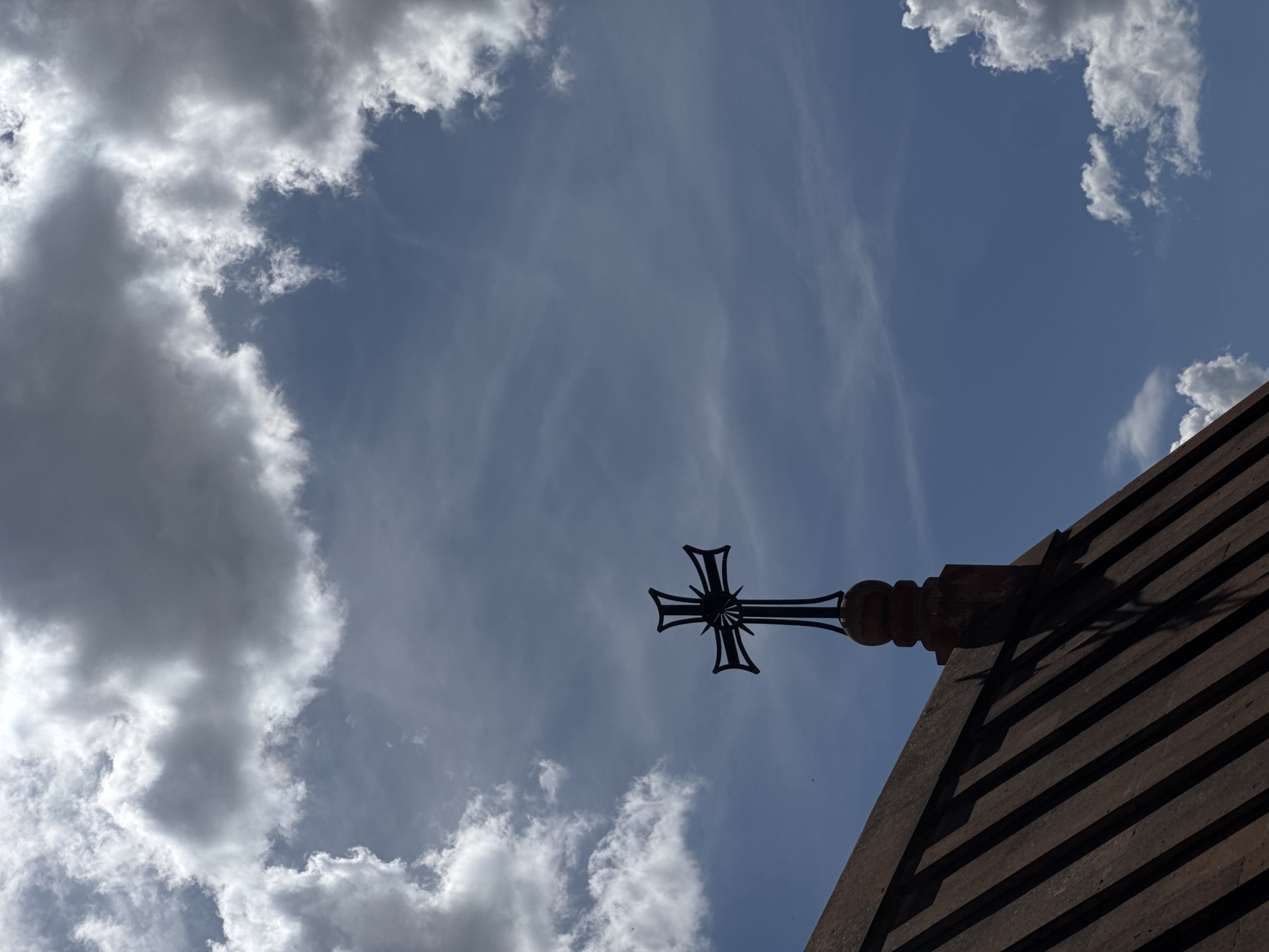 A low-angle shot of the Holy Mother of God Church in Arinj, Armenia shows a black cross affixed to the church roof, set against a cloudy blue sky. The roof is constructed with ribbed, dark-colored material. Light is inconsistent throughout the image.