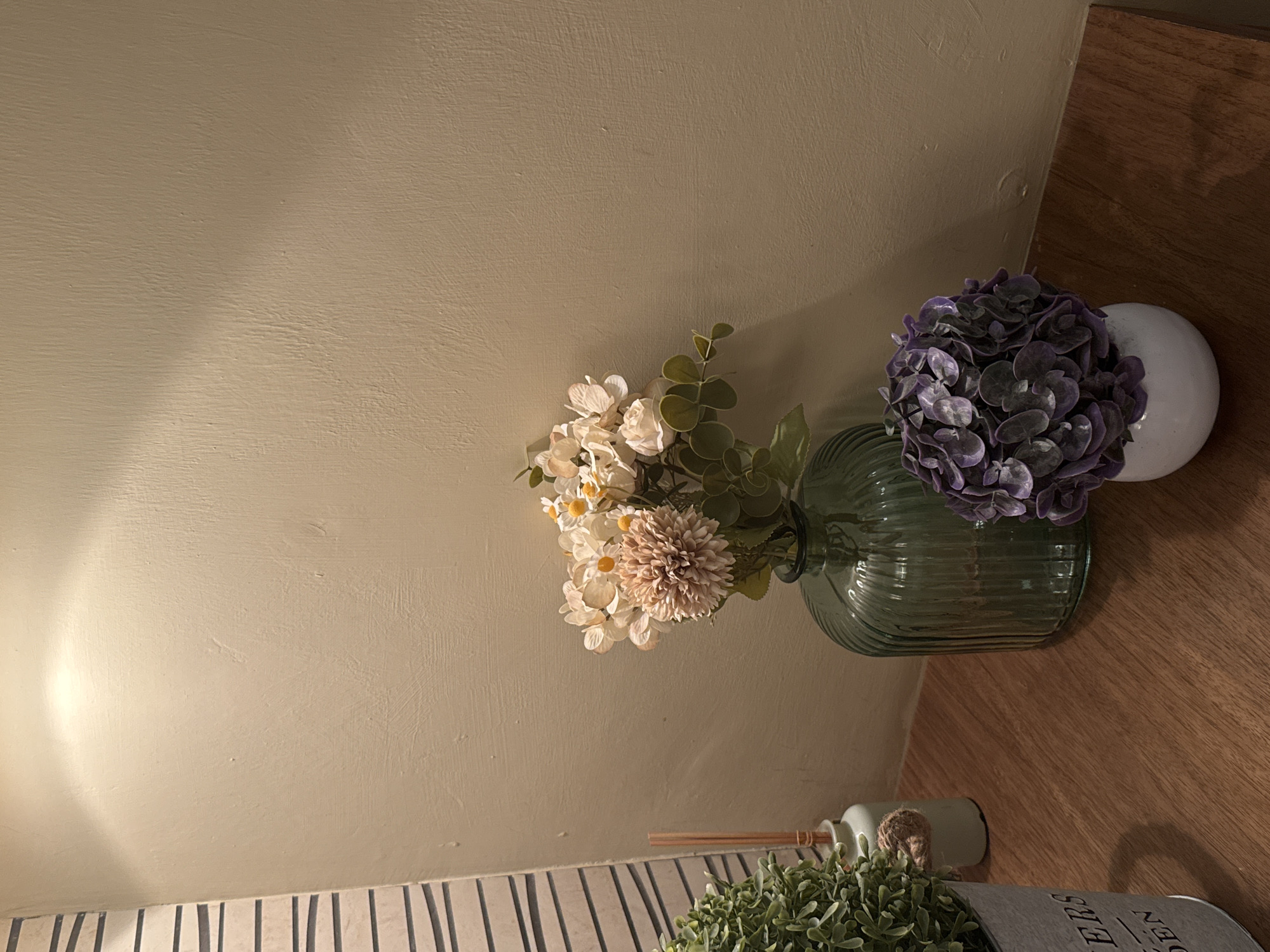 The image shows a still life arrangement against a textured, light beige wall. Situated on a wooden surface, possibly a shelf or table, is a collection of decorative items. There is a green glass vase containing artificial flowers in shades of white, cream, and brown, accented with greenery. Adjacent to this vase is a white orb-shaped vase holding purple artificial flowers resembling hydrangeas. In the lower portion of the frame, there is a small, round, light green bottle with diffuser reeds and a potted artificial plant with small green leaves. The foreground features a striped surface, potentially a tablecloth or part of the furniture. This photo may be found in San Bernardino Tlaxcalancingo, Mexico.