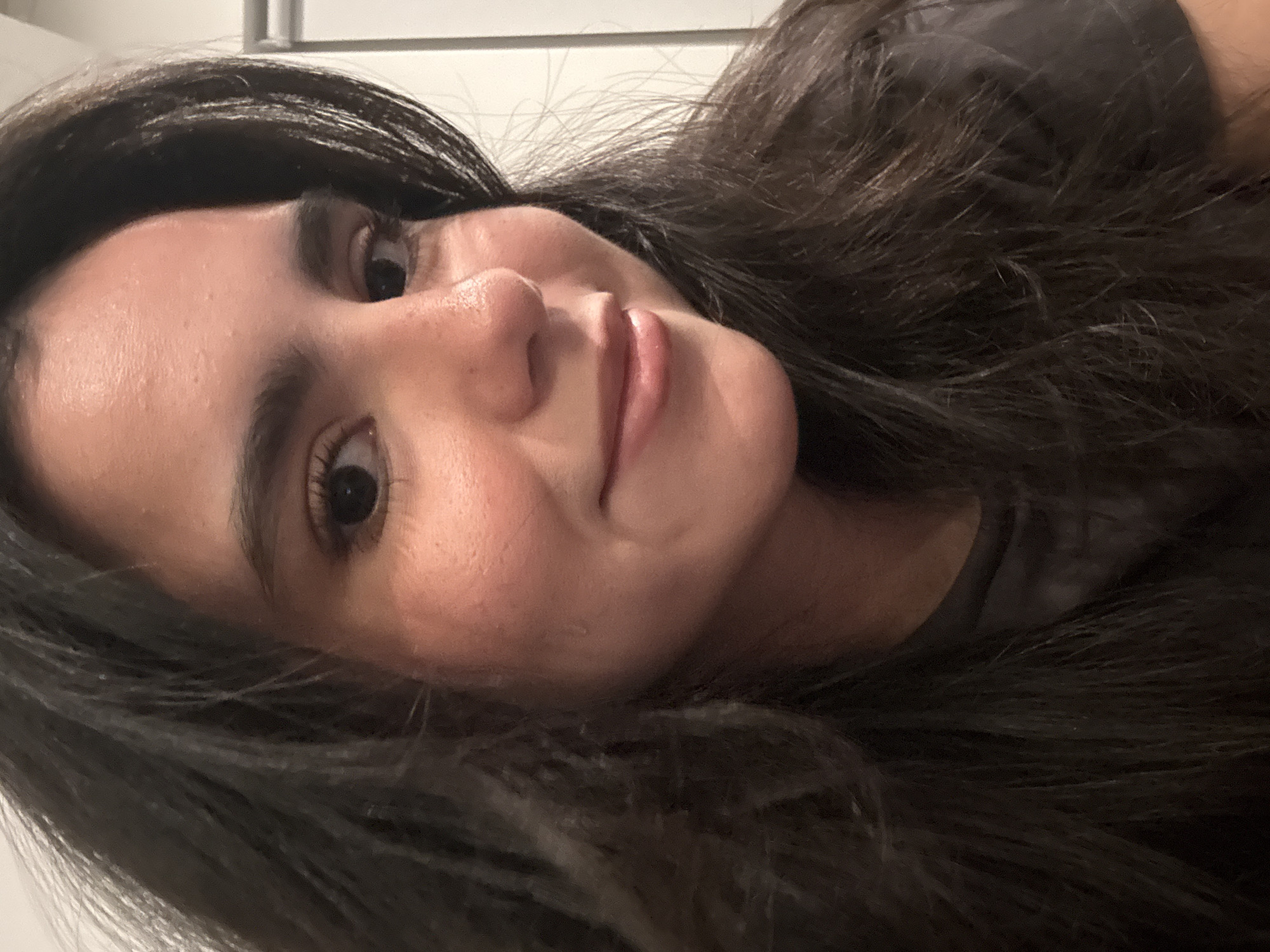 A close-up, slightly angled shot depicts a woman with long, dark hair. Her skin tone appears fair, with subtle rosy hues on her cheeks. She has dark eyes and thick eyebrows. Her lips are slightly parted, revealing a glimpse of her upper teeth. The background is nondescript, with a hint of a white wall or surface visible in the upper left corner. The setting is not readily identifiable as being in Austin, Texas, based solely on the image.