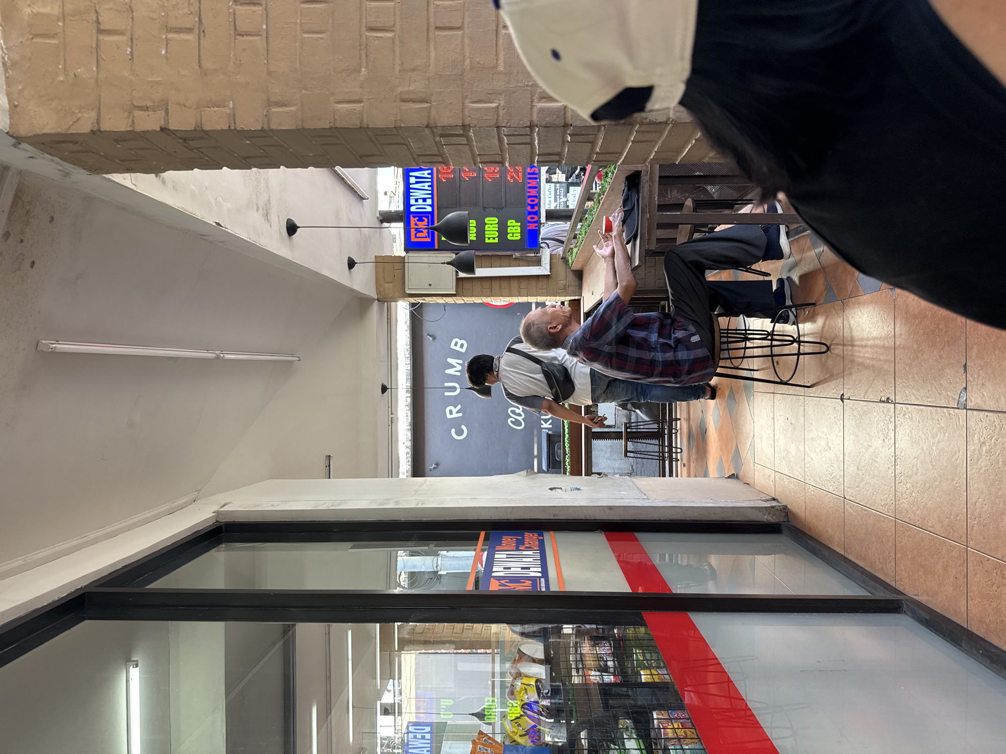 The image depicts a scene from Kuta, Indonesia, showcasing an outdoor corridor with a business named "DNC DEWATA Money Changer" on the left side of the frame. A man wearing a plaid shirt and dark pants is seated at a small metal table with a laptop, seemingly in the middle of an interaction as he holds his hands up. Another person is wearing a white shirt and baseball hat walking the other direction. The corridor is under a brick overhead structure, with tiled flooring. The "DNC DEWATA" signage displays currency exchange rates for EURO and GBP (British Pound) with the phrase "NO COMMISSIONS." The environment suggests a commercial or tourist area.