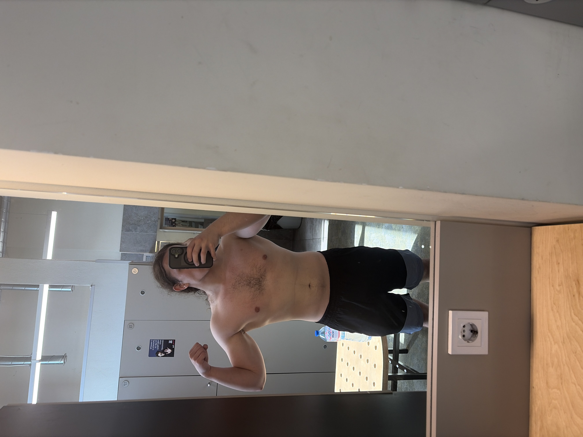 A person with long brown hair, wearing black shorts, is posing shirtless for a mirror selfie in a room in Plovdiv, Bulgaria. The room appears to be a changing room or gym locker area. Behind the person are rows of lockers with numbered compartments, with posters on some of the lockers. A water bottle sits on a light-colored chair. A white electrical outlet is visible on the wall to the right. The ceiling and walls appear to be painted white.