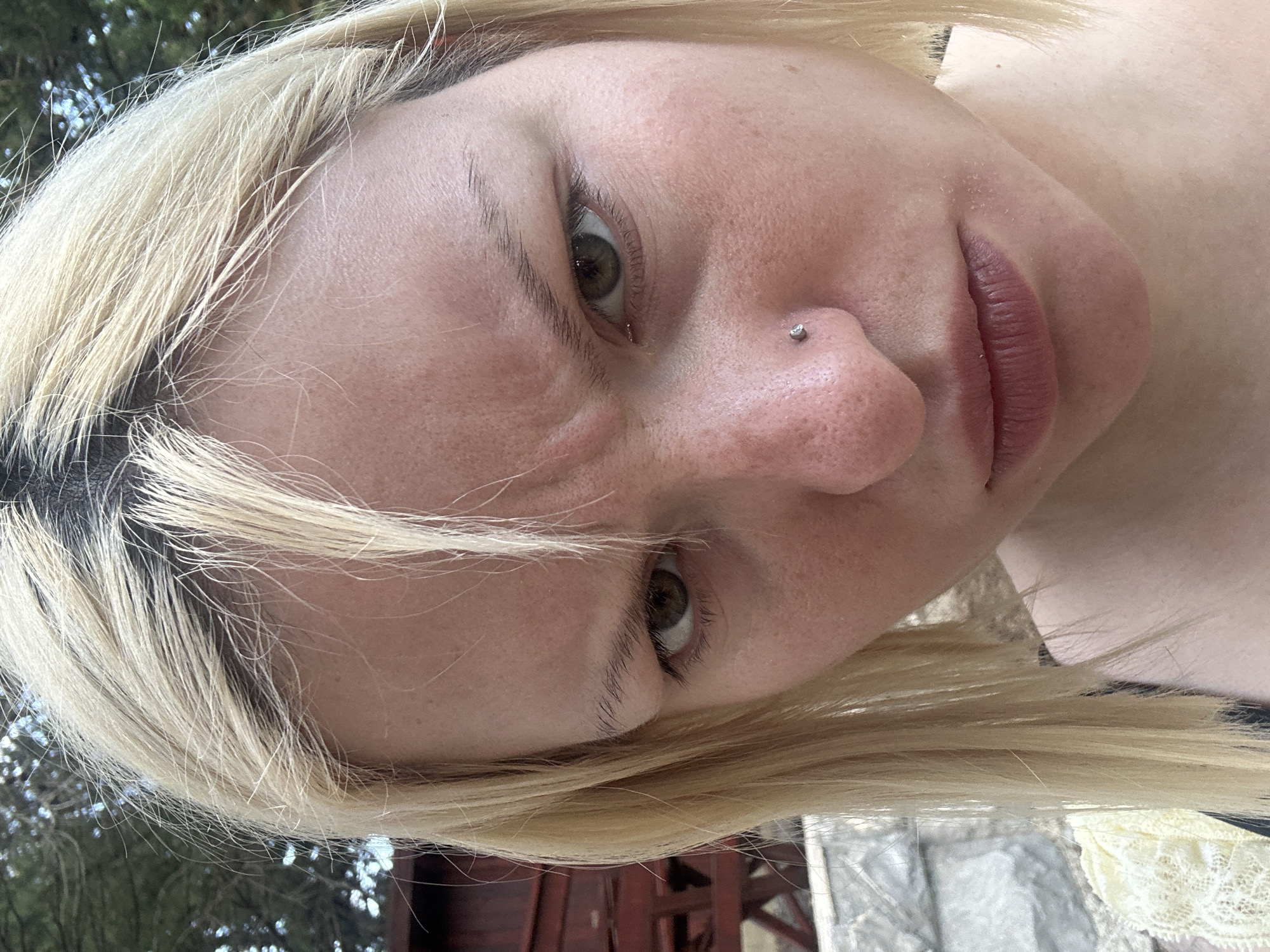 A close-up, slightly angled self-portrait of a blonde woman with fair skin and brown eyes. A silver nose piercing is visible on her left nostril. She appears to be outdoors, possibly in Boreti, Montenegro, as there is foliage and what appears to be a building in the background. The scene is captured in daylight.