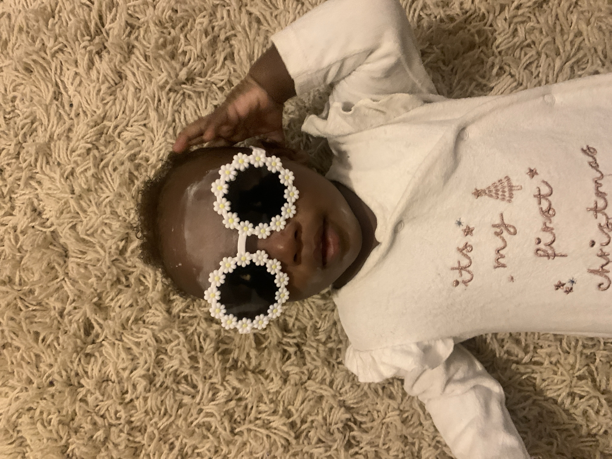 A baby is lying on a tan shag carpet wearing a white onesie that says "It's my first Christmas" in pink script with a Christmas tree design. The baby is also wearing round sunglasses with white daisy frames. The baby's left hand is touching their forehead. The image shows the baby from above. There is no indication of this image being taken in Barakin Akawo, Nigeria.