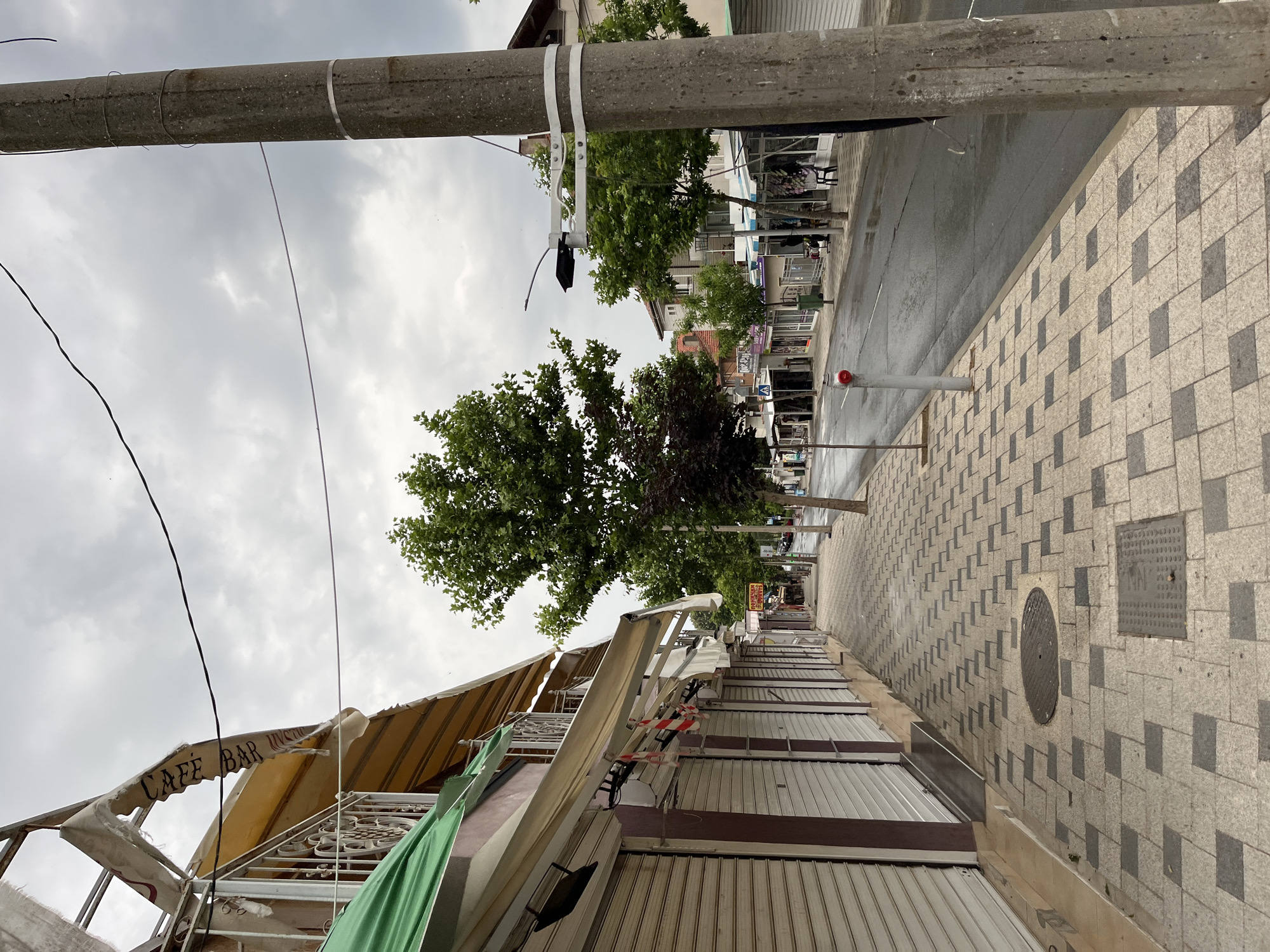 This is a daytime, eye-level, medium shot of a deserted street in Obzor, Bulgaria, under a cloudy sky. The street is lined with closed shops, their metal shutters pulled down. The sidewalk is paved with light and dark grey square tiles arranged in a checkerboard pattern. A single concrete utility pole stands on the right side of the frame, with wires attached to it. Trees with green foliage are visible along the street and in the background. In the distance, more shops and buildings can be seen, along with parked cars. The street appears to be wet, suggesting recent rain. There are no people visible in the frame.