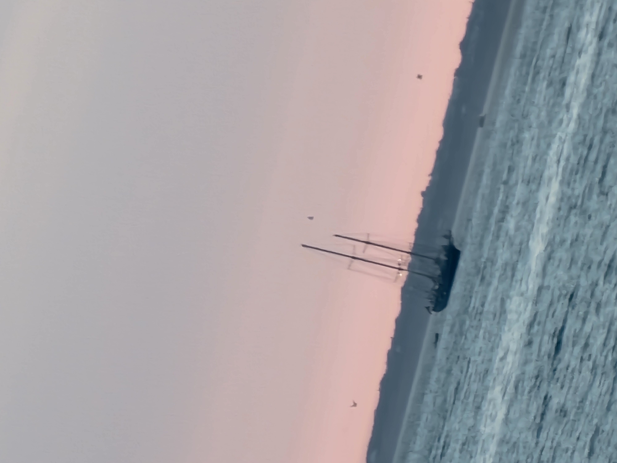 A silhouette of a sailboat with multiple masts is visible on the water, tilted to the right. The sky is a gradient of soft pink and pale grey, suggesting either dawn or dusk. A dark landmass with a subtle, undulating silhouette forms the horizon line. The water is a muted blue-grey, with visible ripples and some lighter foam or wave crests. Several small, dark shapes, possibly birds or distant objects, are scattered in the sky and near the horizon. The overall scene is tranquil and somewhat distant, with the primary subject, the sailboat, being the most prominent object. There are no visible people. The image is slightly blurred and has a tilt, giving it a dynamic or artistic quality. The location is Quarteira, Portugal.