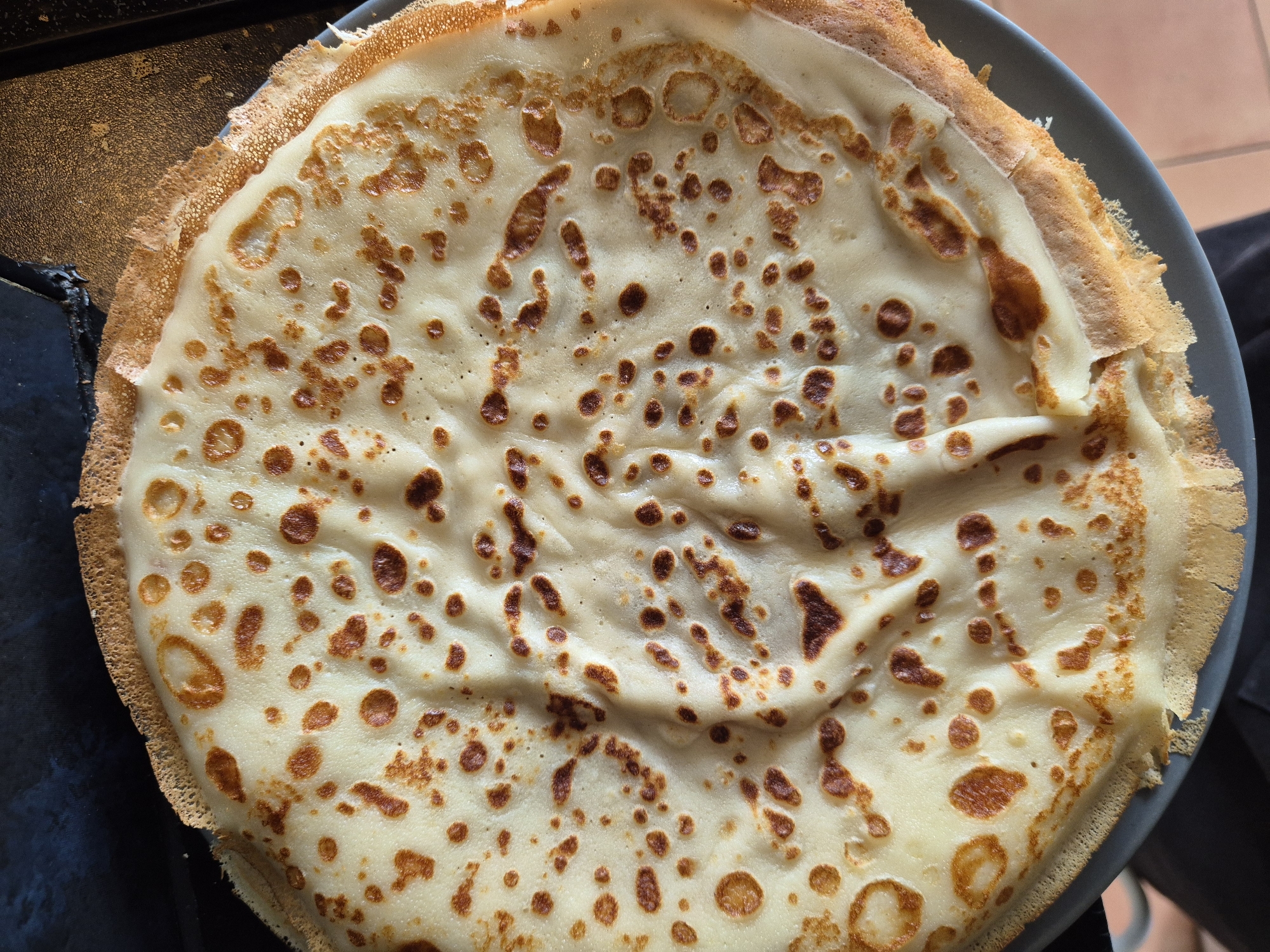 An overhead, close-up photograph displays a stack of at least three circular, thin pancakes or crepes resting on a dark grey plate. The topmost crepe is predominantly a pale cream color, marked with numerous irregular, medium to dark brown caramelized spots across its surface. The texture appears slightly undulating with fine wrinkles. The outer edge of the top crepe is crisp, thin, and displays a more uniform golden-brown hue, with some areas exhibiting a slightly ruffled or torn appearance. The dark grey plate is partially visible along the lower left and right perimeters of the stacked food. In the background, to the upper left, a dark, matte, and subtly textured surface is present. To the upper right, a dark, somewhat reflective surface with visible texture, consistent with a stovetop or pan, is depicted. The lower right background reveals a section of light brown tiled flooring, while the lower left shows a glimpse of dark, soft fabric. No individuals, specific actions, or interactions are visible. The image contains no identifiable information regarding its geographical location, including the city of West Suffolk, United Kingdom.