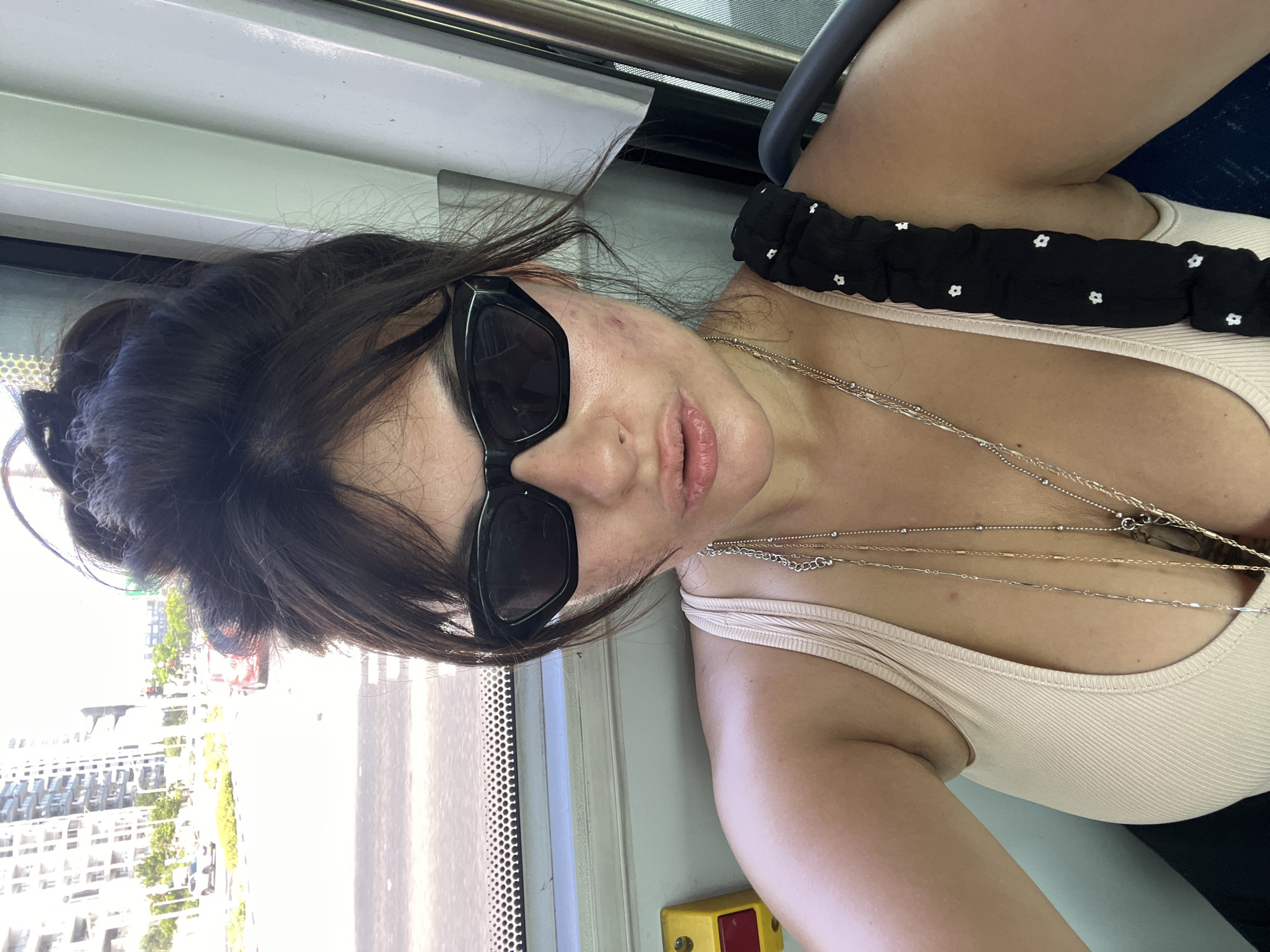 A woman with her hair pulled up in a bun and wearing sunglasses and a light-colored tank top is taking a selfie. She is indoors, possibly on public transportation, as suggested by the window and the yellow emergency button visible on the left. In the background, through the window, a street scene with cars and buildings can be seen, indicating an urban environment. The architecture suggests it could be Kiryat Motzkin, Israel, known for its modern buildings. She is wearing multiple necklaces.