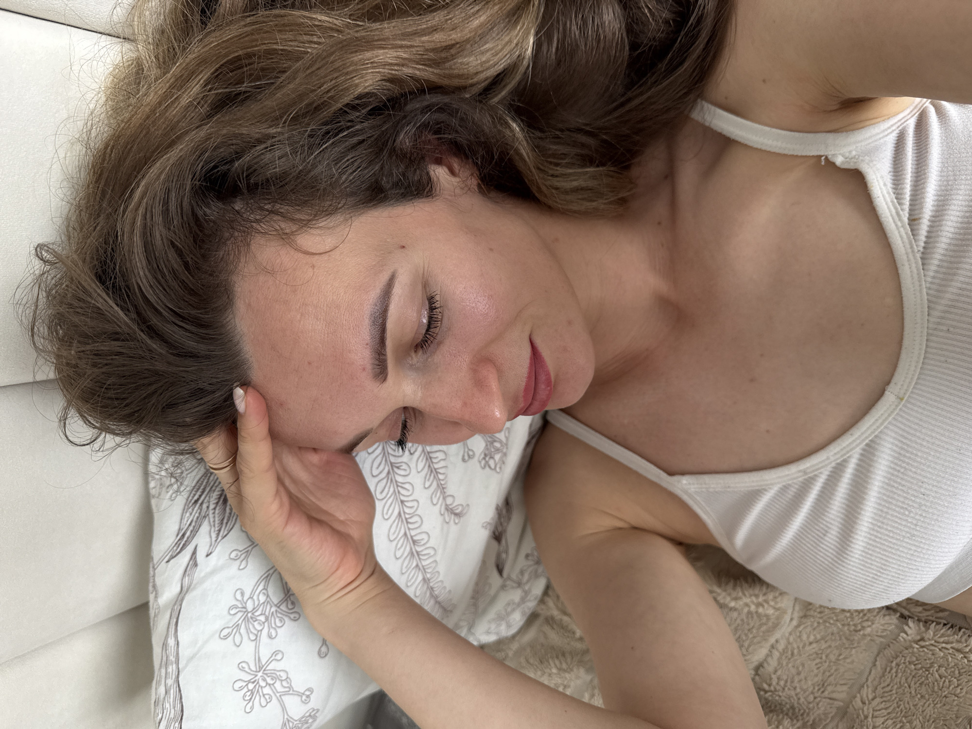 A woman is lying on a bed, looking down with her eyes closed. Her right hand is resting on her forehead. She is wearing a white ribbed tank top. The background consists of a pillow with a floral pattern and a textured blanket. The scene is lit by natural light. This image was taken in the city of Perm, Russia.