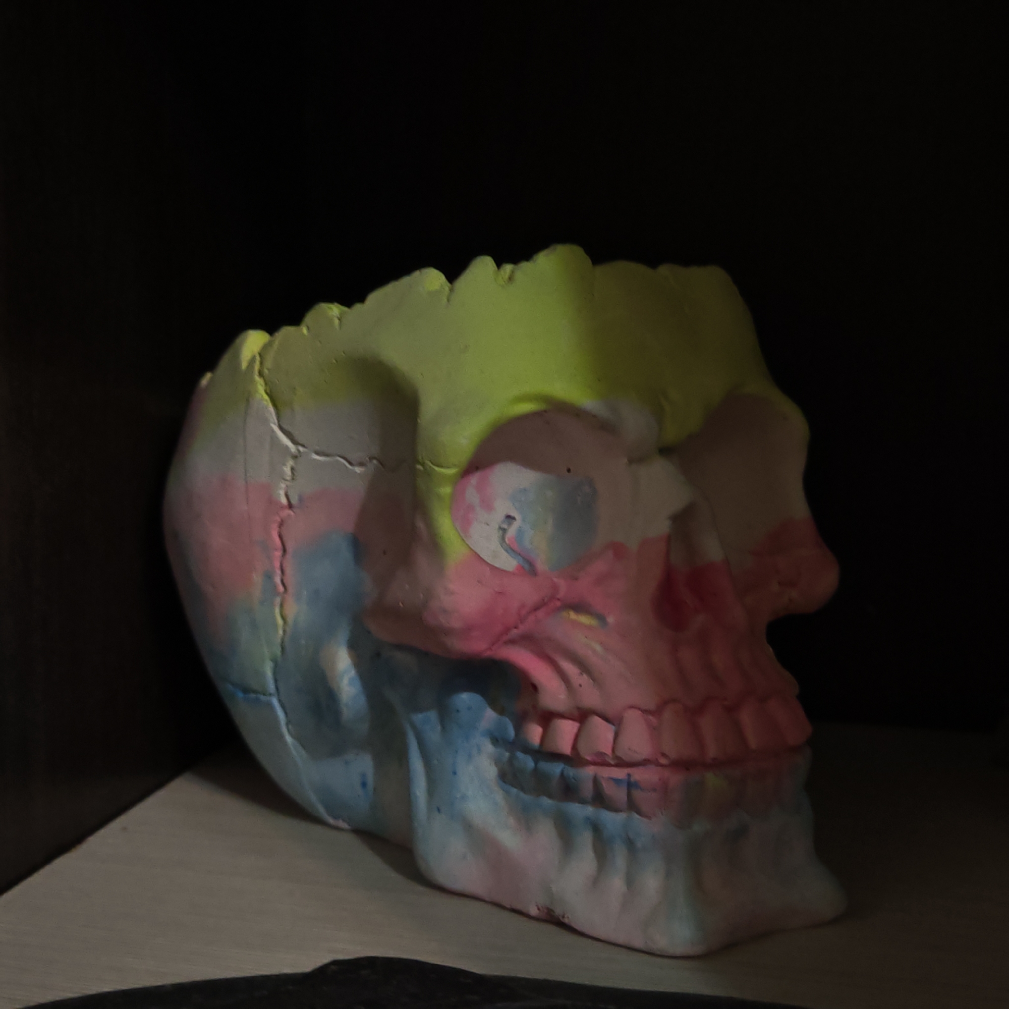 A brightly colored skull sits on a surface. The skull is painted with sections of neon yellow, pink, and blue. It appears to be a decorative object. The background is dark, suggesting the skull is on a shelf or in a dimly lit space. The specific location in Kladovitsy, Russia, is unknown.