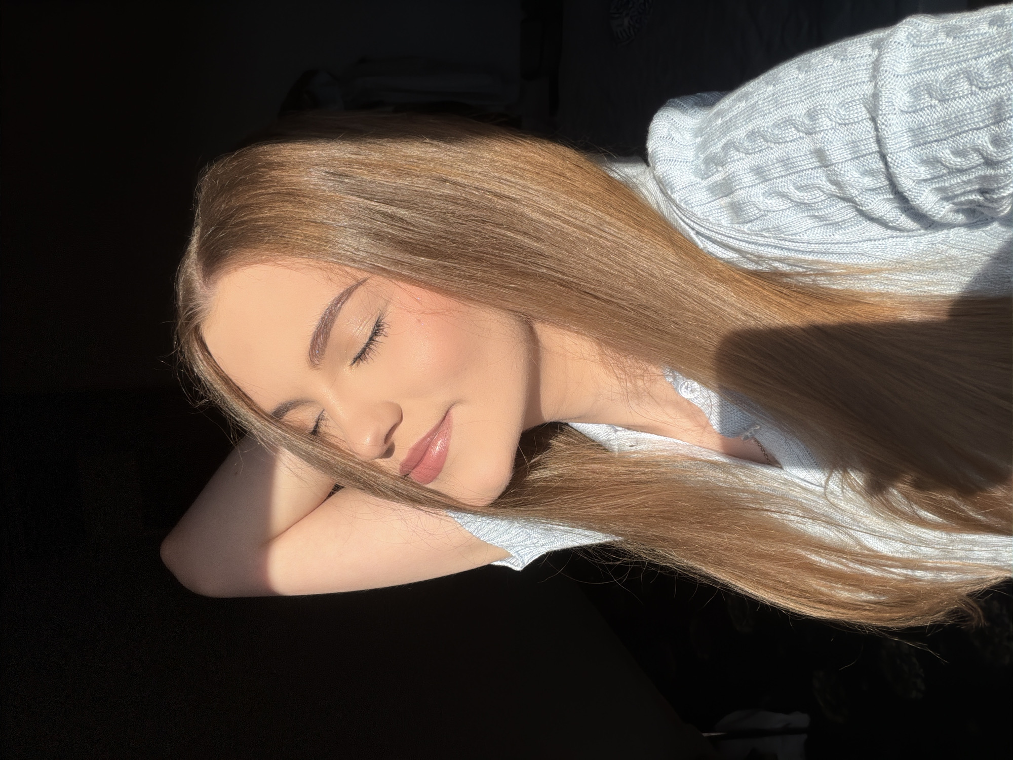 A young woman with light brown hair is photographed from the chest up, indoors, with her eyes closed and her right arm behind her head. Sunlight illuminates the right side of her face and hair, casting a shadow on the left. She is wearing a light blue cable-knit sweater. The background is dark, with some indistinct shapes of furniture visible on the right. The image is taken in a domestic setting in Dnipro, Ukraine.