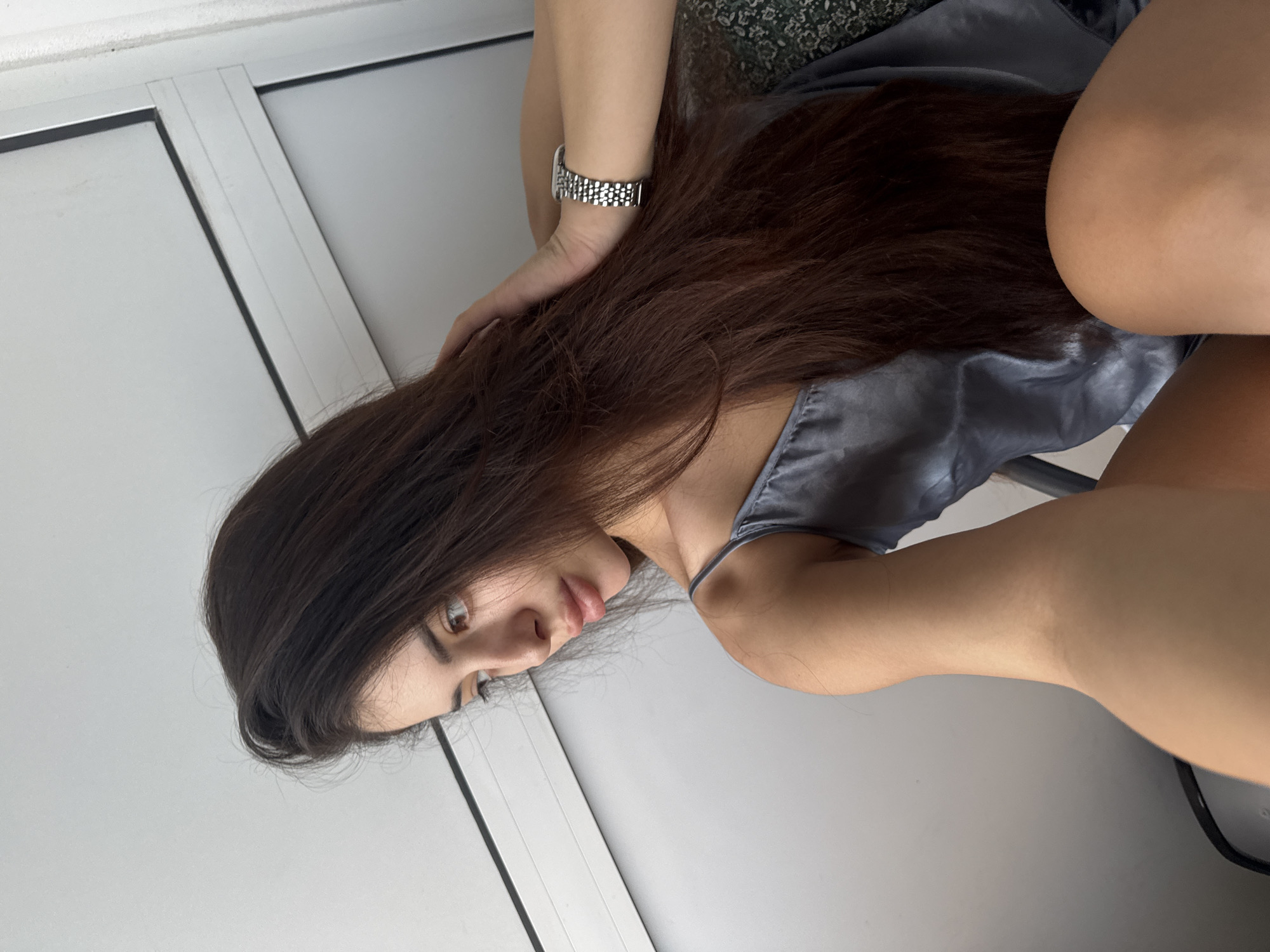 A young woman is captured in a medium close-up, turned sideways with her head tilted downwards, looking off-frame. She has long, flowing dark brown hair that cascades over her left shoulder and arm. Her complexion is fair, and her expression appears neutral or contemplative. She is wearing a light blue-grey camisole or slip-style top with thin straps, made of a satin-like material. A silver-toned watch with a metal link bracelet is visible on her left wrist.
Her left arm is bent, with her hand touching the back of her head/hair. Her right arm is also bent, and part of her leg or upper thigh is visible in the lower right portion of the frame. She appears to be in a relaxed, possibly lounging, pose.
The background suggests an indoor setting, likely in Plovdiv, Bulgaria. To her left, there are light grey or white vertical panels, possibly part of a window frame or a sliding door, separated by a silver metallic strip. Behind her head and shoulder, a dark patterned fabric, possibly part of a cushion or furniture upholstery, is visible with a subtle floral or leaf design. The lighting is soft and even, indicating diffused natural light or gentle artificial illumination. No visible text is present in the image.