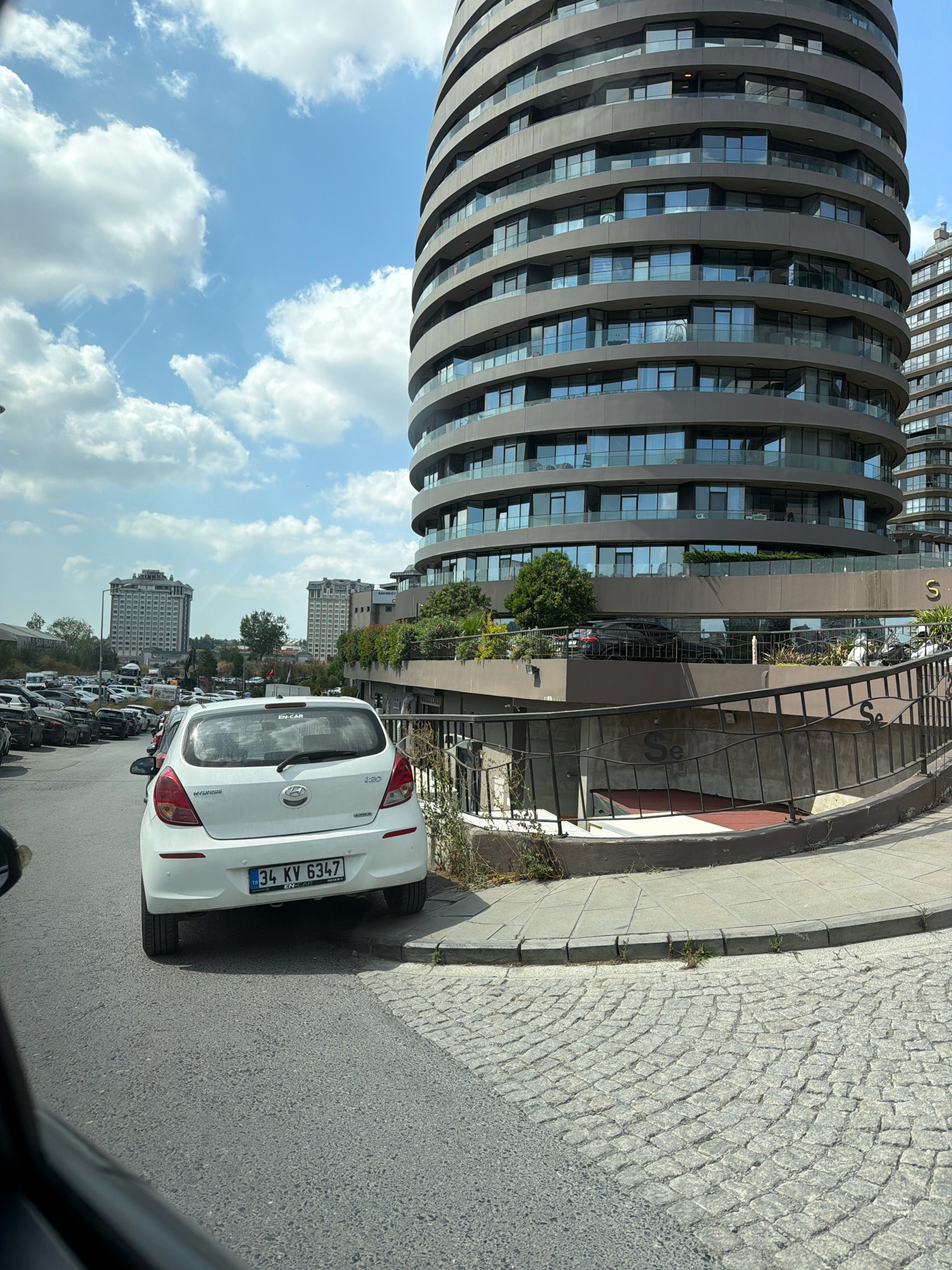 This is a street-level, eye-level, medium-shot photograph of a white Hyundai i20 vehicle parked on the right side of a paved road. The car is facing away from the camera and its license plate reads "34 KV 6347". To the right of the car, a sidewalk made of cobblestones leads to a modern, multi-story building with a curved facade. This building has many floors with windows and balconies. In the background, other buildings and a cloudy blue sky can be seen. The scene appears to be a street in Bakırköy, Turkey, with the curved building likely being part of a residential or commercial complex. There are no people visible in the immediate vicinity of the car. The road surface is asphalt, and there is some vegetation along the sidewalk.