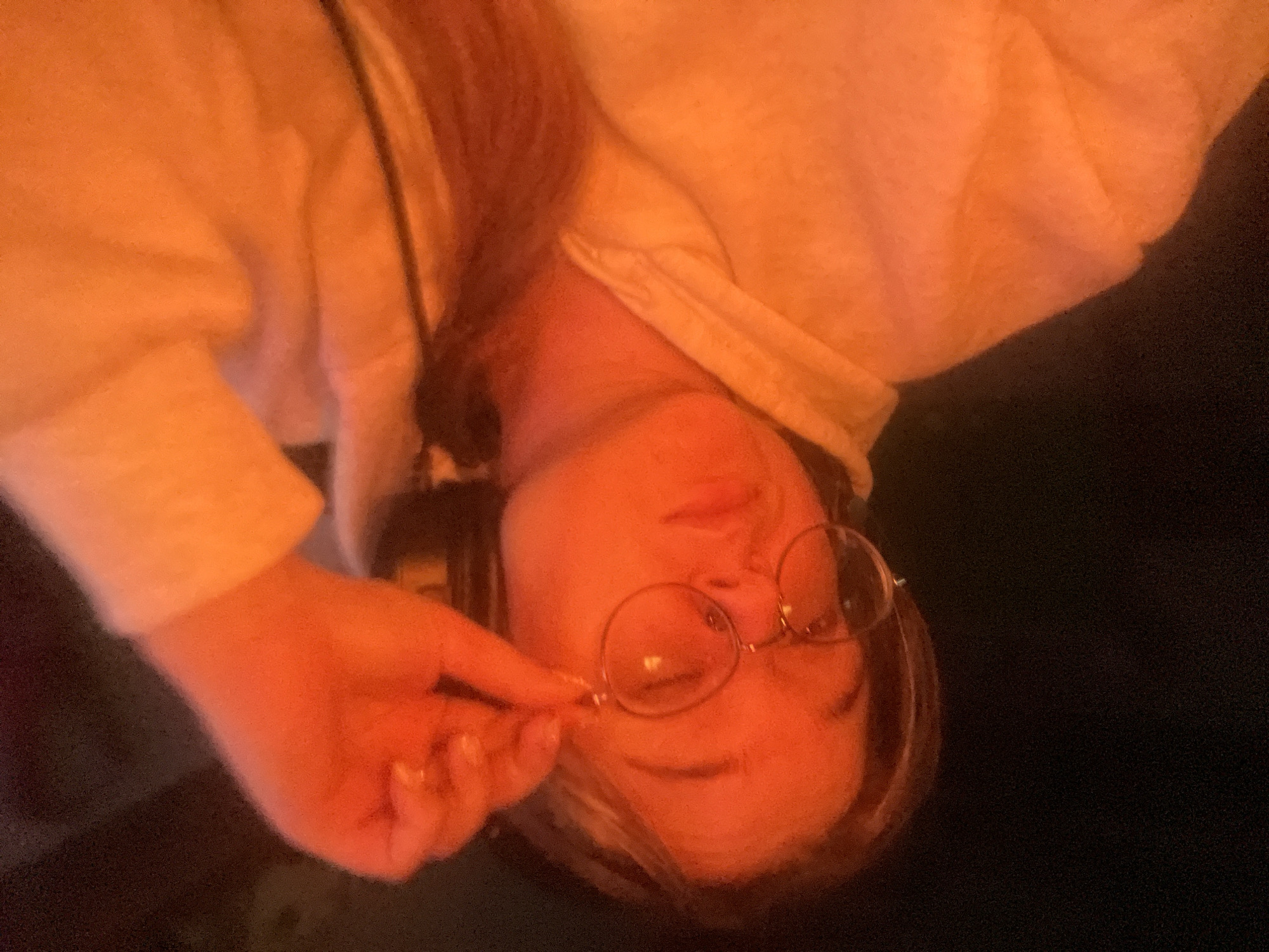 A young person with blonde hair is shown from the chest up, wearing glasses and headphones. They are adjusting their glasses with their right hand. The person is wearing a light-colored hooded sweatshirt. The lighting is predominantly orange, suggesting an artificial light source or a time of day with warm lighting. The background is dark and out of focus. There is no information about the location in the city of Kolomna, Russia.