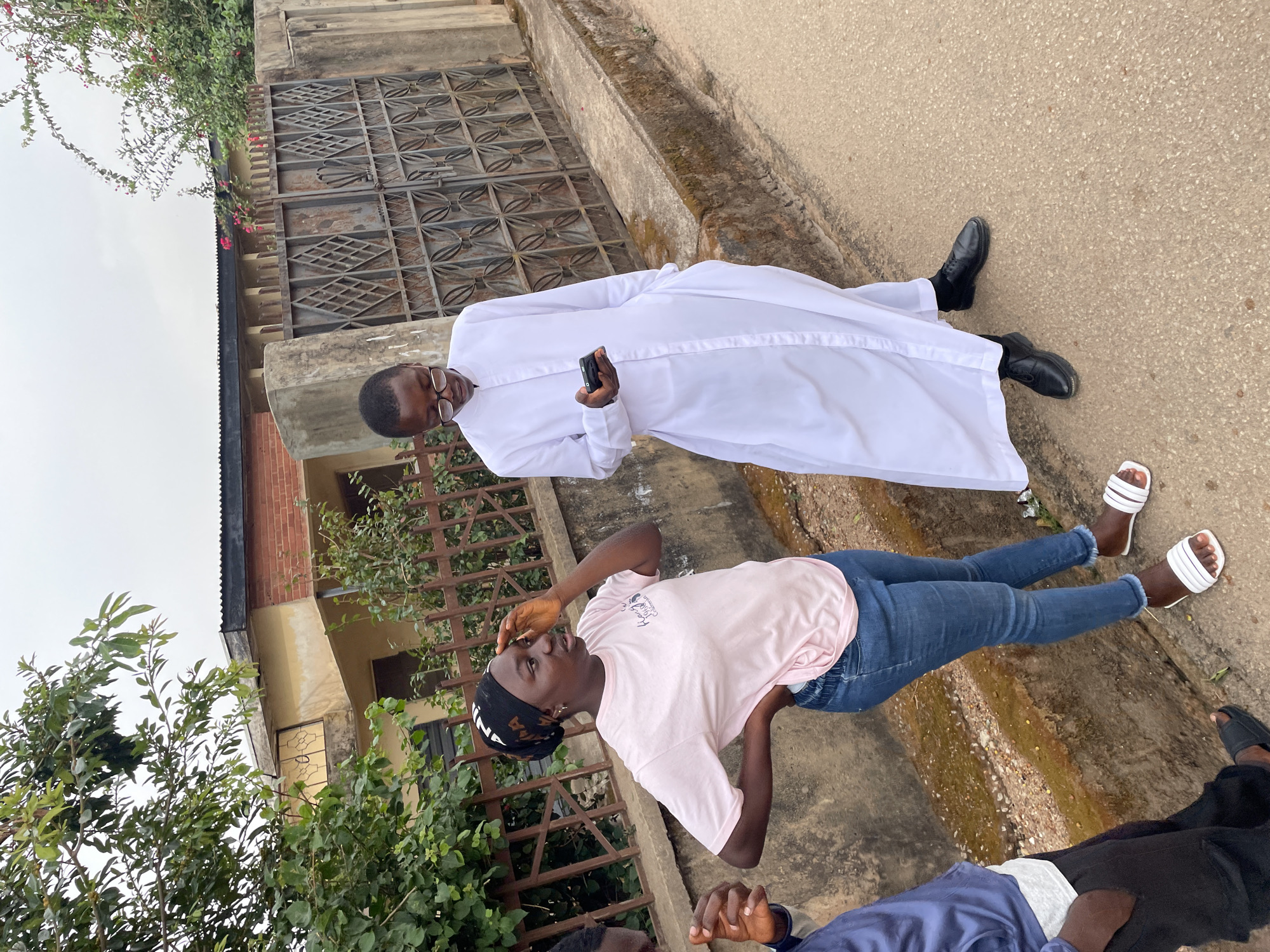The image depicts three individuals outdoors on a gray concrete sidewalk in Jos, Nigeria. An adult male in a long white robe and black dress shoes stands facing left, holding a dark-colored mobile phone. A younger person wearing a pink t-shirt, blue jeans, and white sandals is positioned perpendicular to the first person, facing towards the camera with their hand on their forehead. A third individual is partially visible at the bottom edge of the frame, wearing a blue shirt. Green vegetation and a gray metal gate are visible on the left, indicating a residential or commercial setting. The sky appears overcast. The buildings in the background show some wear and tear, consistent with an urban environment.