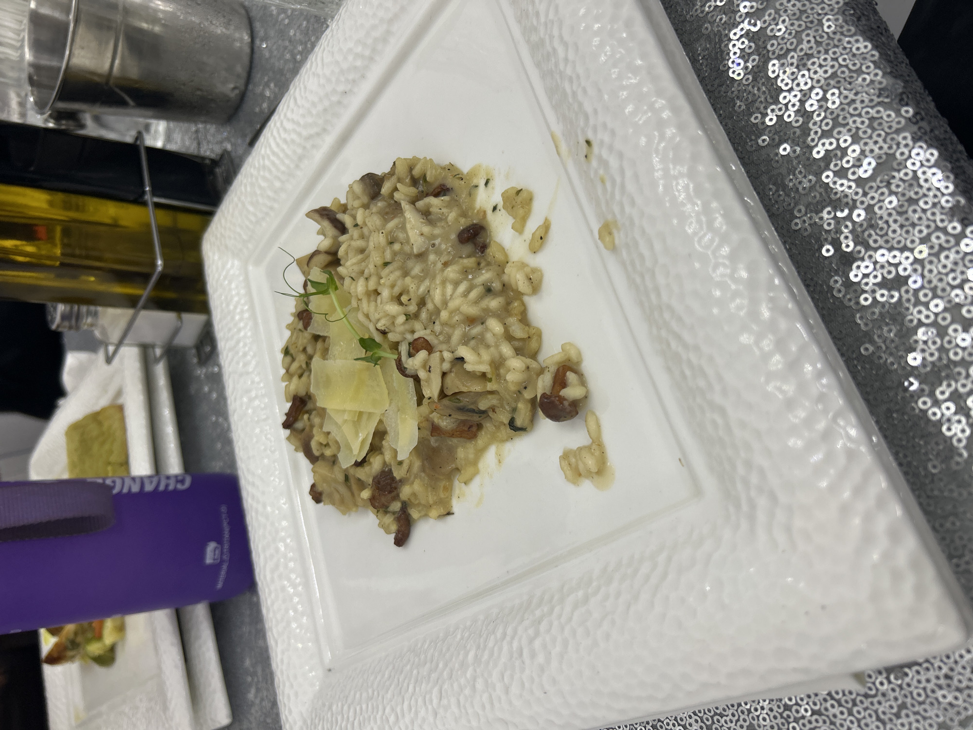 The image shows a white square plate with textured edges, containing risotto with mushrooms and a garnish of Parmesan cheese and microgreens. The plate sits on a sparkly, sequined tablecloth. To the left of the plate, there's a purple object with the word "CHANG" printed on it, along with other dishes and serving ware in the background. The setting appears to be a restaurant, though the specific location in Plovdiv, Bulgaria, cannot be determined from the image alone. There are no people visible in the frame.