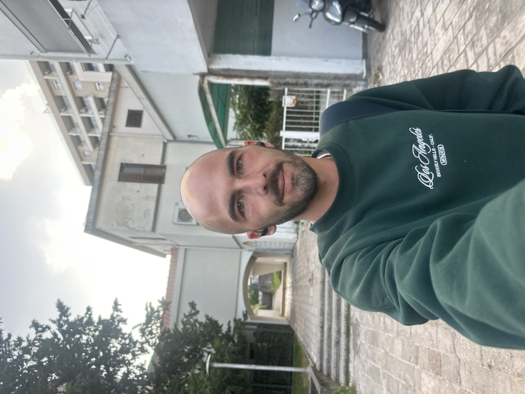 A man with a shaved head and beard is the central subject of the photo, wearing a green "Los Angeles" sweatshirt. He is positioned at an angle, suggesting he might be holding the camera for a selfie. He has earbuds in. The background reveals urban scenery, including a multi-story building to the left and a motorcycle parked near a building with green shutters. A tree is visible on the bottom left of the image. The ground is paved with grey bricks. The picture appears to have been taken in Budva, Montenegro, as the architectural style and vegetation are characteristic of the region.