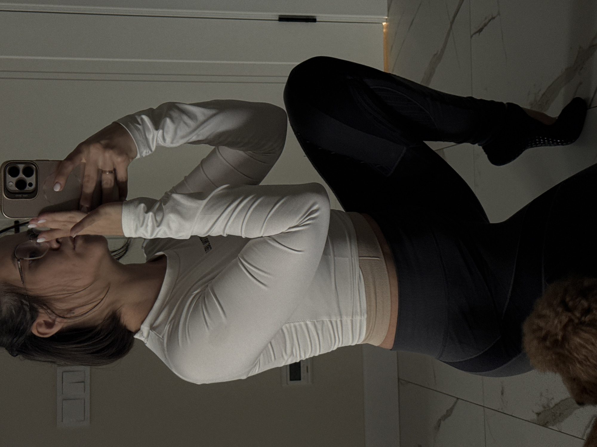 In a mirror selfie taken indoors, a person is seen in a relaxed, seated or kneeling position on a light-tiled floor. They are holding a rose gold/gold iPhone, featuring a triple camera system, with both hands, appearing to look down at the screen. A simple band ring is visible on the ring finger of their left hand.
The person wears a white long-sleeve top, which has faint, unreadable text on the chest. A light beige or nude-colored ribbed waistband is visible around their midsection, above dark gray or black athletic leggings with subtle textured panels on the sides. Their feet are covered by dark, sock-like footwear with visible grippy dots on the soles.
The background consists of light-colored walls, possibly a door frame or baseboard visible behind the person's head and arms. The floor has a light-colored, marble-like tile pattern. The overall lighting is dim, casting soft shadows. Part of a furry brown object, possibly a pet or a piece of decor, is visible in the bottom right corner of the frame. No specific readable text is clearly discernible on the phone or clothing.
