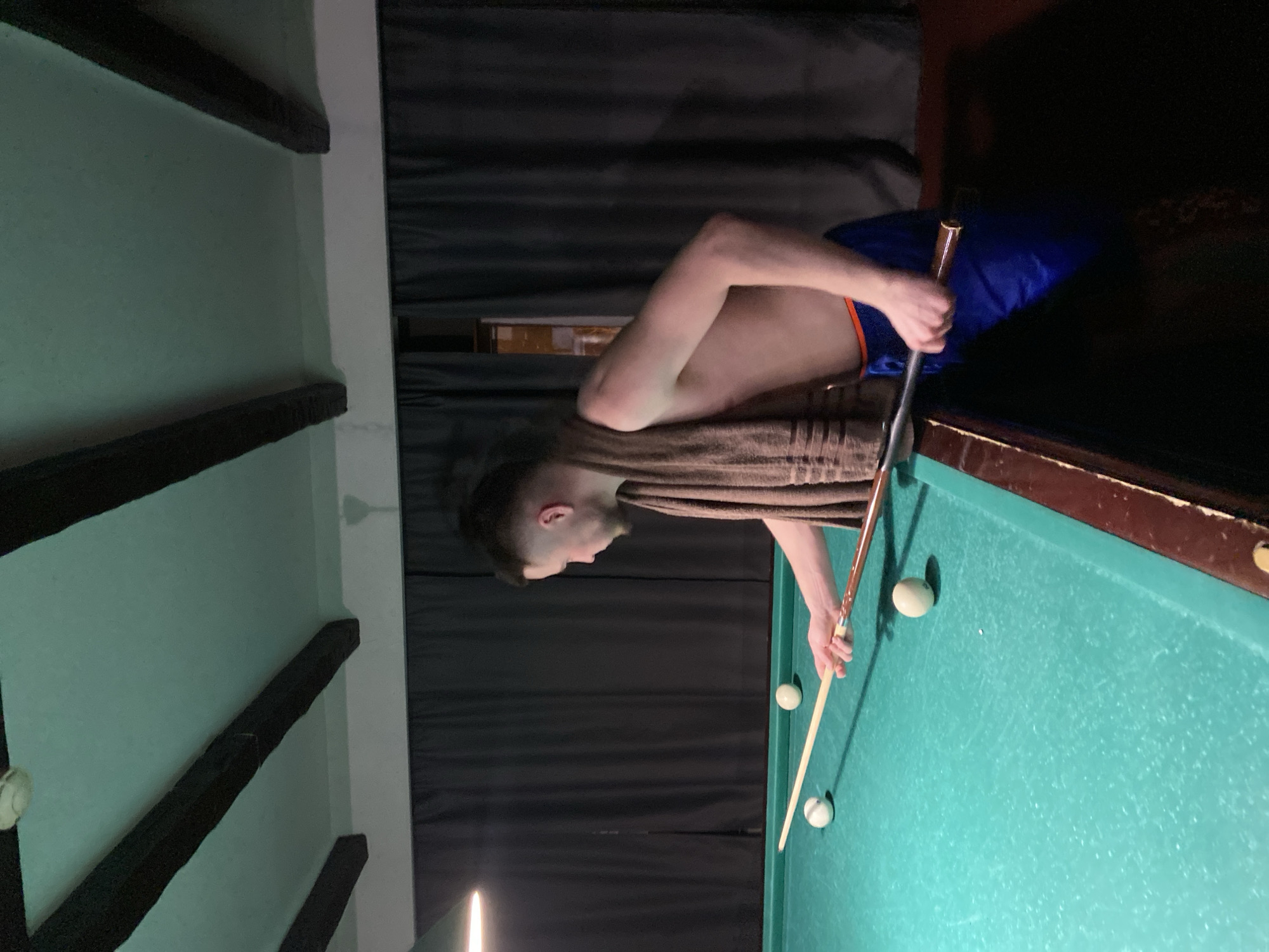 An indoor scene depicts a male individual engaged in playing billiards. The specific location within Perm, Russia, is not discernible from the image. The man, appearing to be in his mid-20s to early 30s with short dark hair and light facial stubble, stands shirtless with a brown striped towel draped over his left shoulder. He wears blue athletic shorts with an visible orange waistband. He is positioned on the right side of a green-felted billiard table, leaning over it, holding a two-piece billiard cue with both hands, poised to strike one of three visible white billiard balls. The green felt of the table exhibits signs of wear. The background features dark grey or black curtains extending across the width of the frame, behind which a faint orange glow is visible, indicative of an exterior light source or window. The ceiling is light-colored, presenting a slight greenish tint, and is characterized by several exposed dark wooden beams running horizontally. A white smoke detector or similar fixture is mounted on the ceiling in the upper-left. An overhead light fixture, partially visible on the upper-left, illuminates the table. The floor in the lower-right foreground is dark.