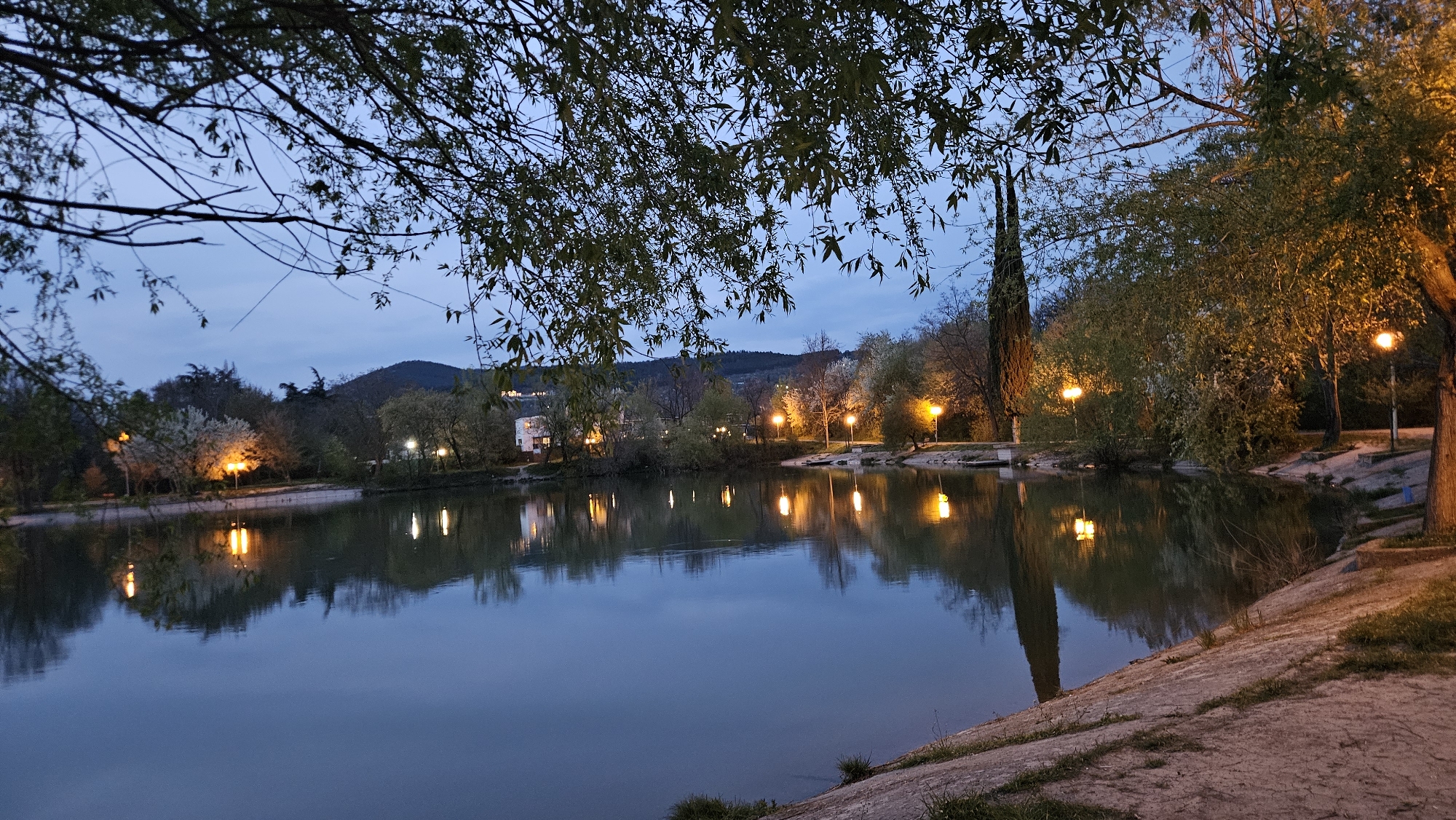 The image depicts a serene lake scene at dusk in the city of Stara Zagora, Bulgaria. The water reflects the soft glow of strategically placed lampposts along the shoreline, creating symmetrical patterns of light and dark. The shoreline is a combination of concrete embankments and natural, uneven ground with trees. On the far side of the lake, buildings can be seen, their forms softened by the low light. A backdrop of rolling hills sits in the distance, visible under the twilight sky. Branches and foliage frame the top of the image, adding depth and framing.