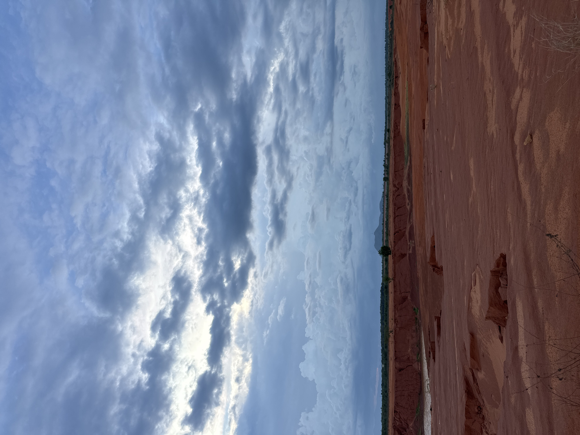 The image displays a vast, arid landscape with striking red rock formations and a dramatic cloudy sky. The foreground is dominated by the textured surface of eroded red earth, showing signs of wind and water weathering, with some dry vegetation clinging to the edges. These formations suggest a desert or semi-arid environment, possibly a canyon or badlands. In the background, a thin line of greenery marks the transition to a flatter, distant horizon. The sky is filled with a dynamic mix of light and dark clouds, creating a sense of depth and atmospheric movement. The clouds range from wispy, scattered formations to darker, more substantial masses, with sunlight breaking through in places, illuminating the scene with a diffused, ethereal light. The overall impression is one of natural beauty and geological grandeur, with the raw power of the elements evident in the sculpted landscape and the ever-changing sky. The time of day appears to be either early morning or late afternoon, given the soft, atmospheric lighting. The scene evokes a sense of isolation and tranquility, inviting contemplation of the natural world's timeless processes.