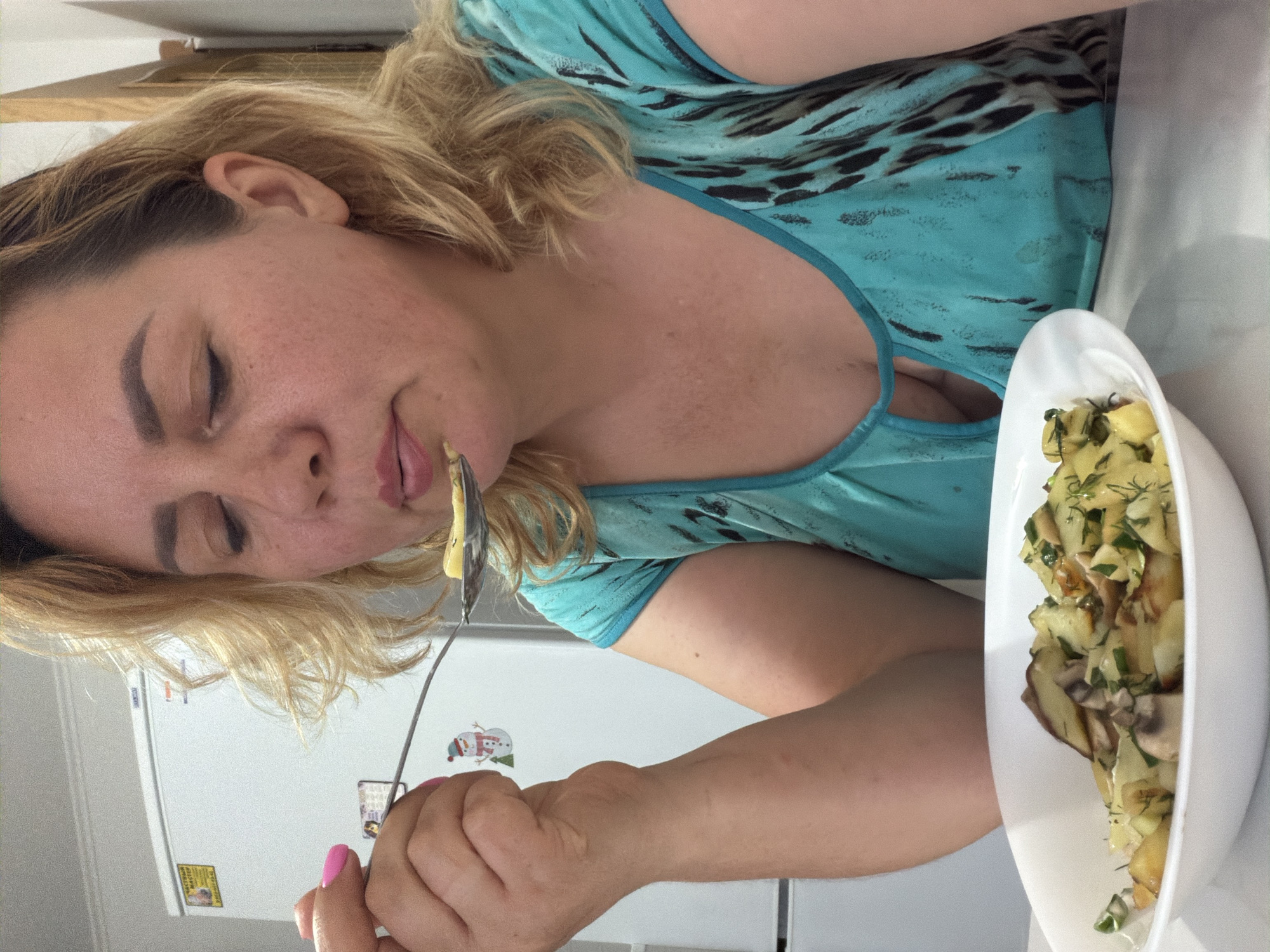 A woman with blonde hair and fair skin is eating from a white bowl with a fork in what appears to be a kitchen. She has a piece of food on the fork and is about to put it in her mouth. The bowl contains what appears to be a potato salad dish with dill. The background reveals a white refrigerator. While the location is indicated as Leningradskaya, Russia, there is no visual evidence in the image to definitively confirm this location.